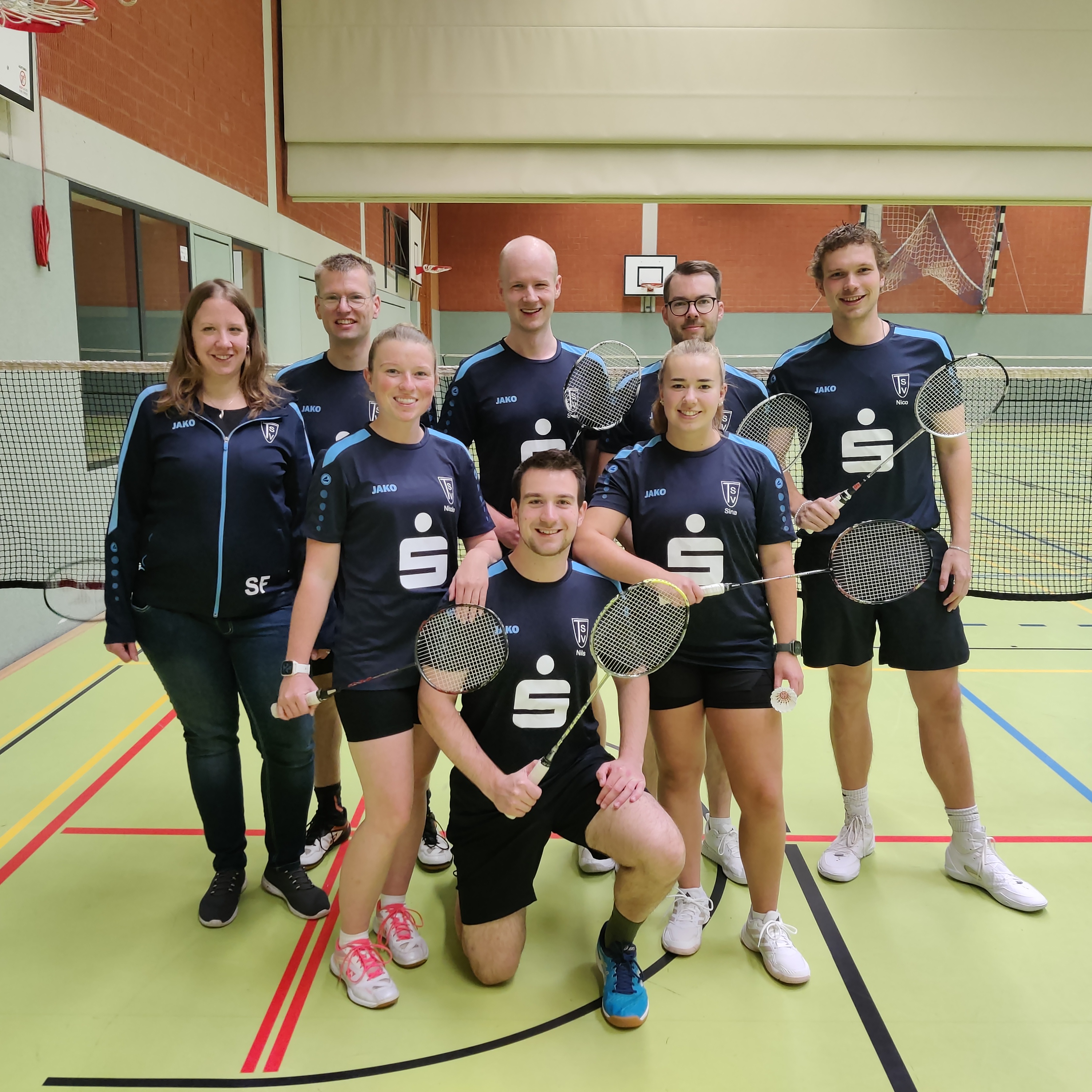 Badminton erster Spieltag 2025_26 Badmintonmannschaft in Sportkleidung mit Schlägern in einer Halle, einige tragen schwarze Trikots mit weißem Logo und Schriftzug, eine Person trägt eine schwarze Jacke mit Initialen 'SE'.