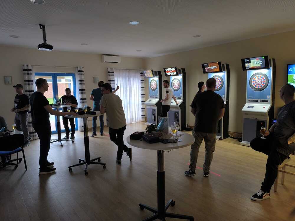 Handicap 1 Mehrere Personen stehen in einem hellen Raum mit Holzfußboden und spielen an elektronischen Dartscheiben, die an der Wand aufgestellt sind. Die Dartscheiben sind in weißen Gehäusen mit Bildschirmen darüber eingebaut. Im Vordergrund stehen runde Stehtische mit Getränken darauf. Die Fenster sind mit gestreiften Vorhängen dekoriert, und an der Decke hängt ein Beamer. Die Atmosphäre wirkt entspannt und gesellig.
