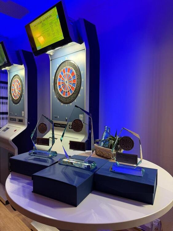 Handicap 2 Drei gläserne Pokale mit eingelassenen Dartboards stehen auf einem runden weißen Tisch. Im Hintergrund befinden sich zwei elektronische Dartscheiben mit beleuchteten Anzeigetafeln, die gelb leuchten. Die Szene ist in blaues Licht getaucht.