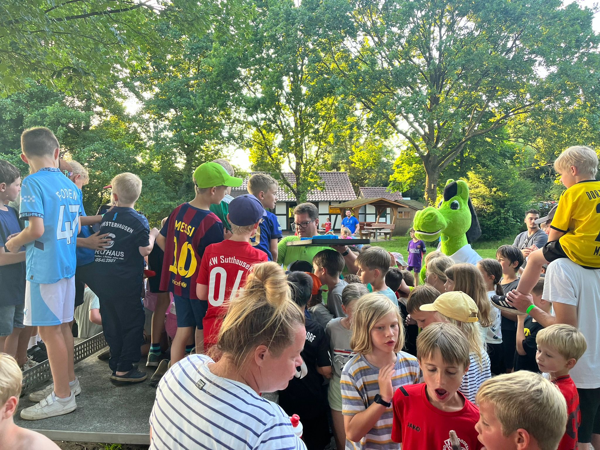 Das Bild zeigt eine große Gruppe von Kindern und einigen Erwachsenen, die sich um ein kleines, erhöhtes Podest versammelt haben. Im Vordergrund stehen viele Kinder, einige tragen Fußballtrikots verschiedener Mannschaften. Im Hintergrund sind ein paar Erwachsene zu sehen, sowie ein grünes Maskottchen, das aussieht wie ein Drache oder eine ähnliche Fantasiefigur. Der Ort scheint ein Außenbereich zu sein, möglicherweise ein Sportplatz oder ein Lager. Die Stimmung wirkt fröhlich und ungezwungen, es scheint ein gemeinschaftliches Event zu sein.
