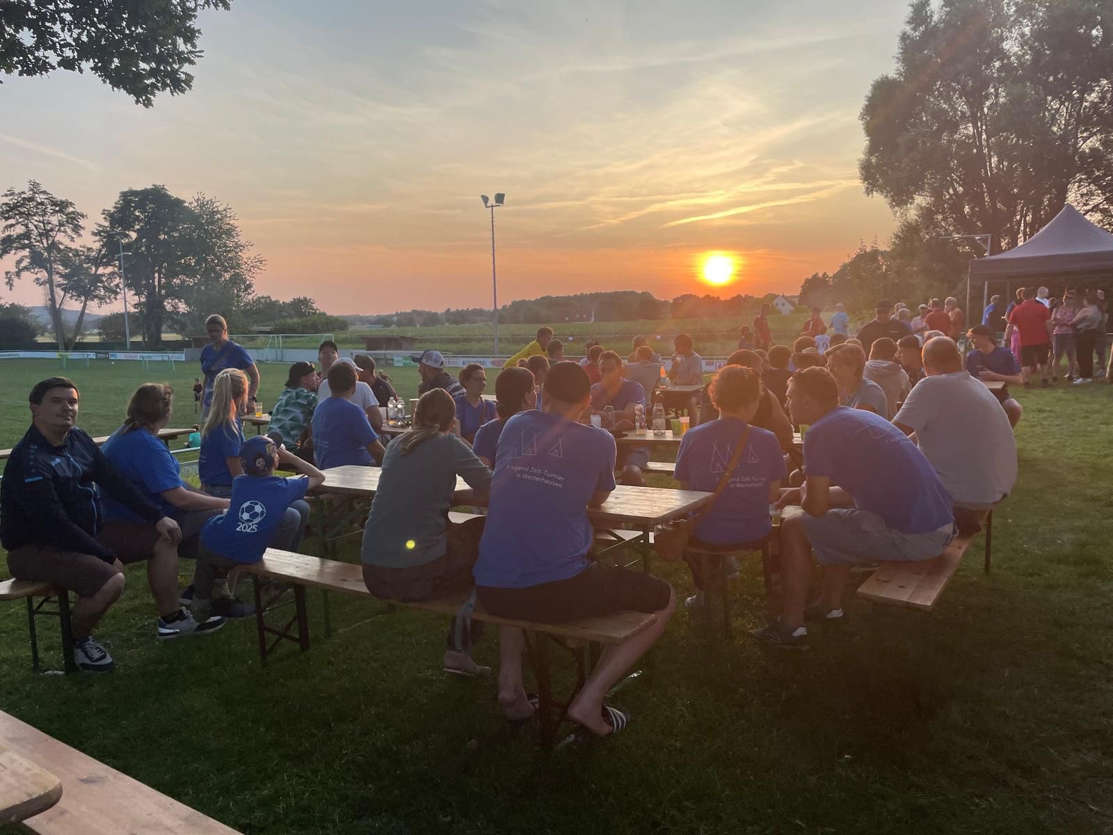 Das Bild zeigt eine große Gruppe von Menschen, die an Biertischen auf einer grünen Wiese sitzen und sich unterhalten. Im Hintergrund ist ein Fußballplatz zu sehen, und die Sonne geht hinter den Bäumen unter, was eine warme, goldene Beleuchtung erzeugt. Die Atmosphäre wirkt entspannt und gesellig, es scheint sich um ein informelles Treffen oder eine Feier im Freien zu handeln. Einige der Personen tragen einheitliche T-Shirts mit einem Logo, was auf ein Team oder eine Organisation hindeuten könnte. Das Bild strahlt eine positive und gemütliche Stimmung aus.