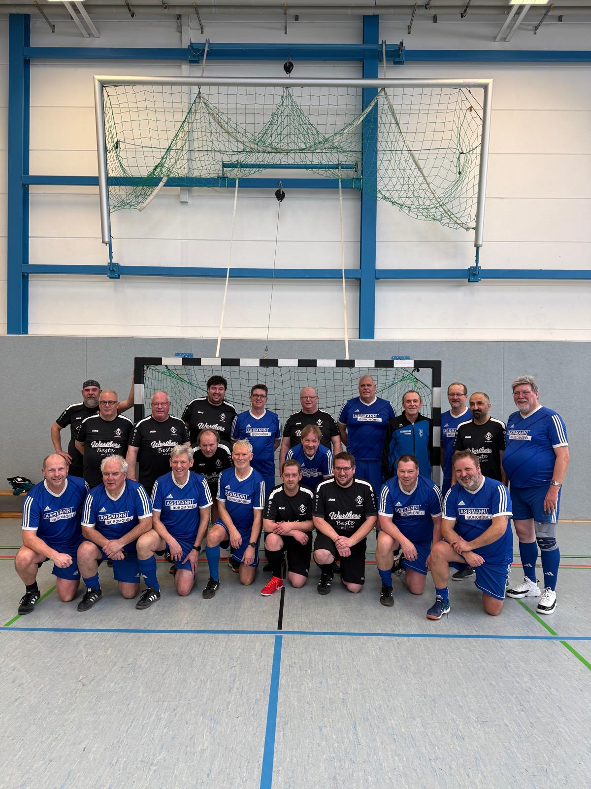 Walking Football mit Testspiel gegen Werther am 08.02.2026 Das Bild zeigt ein Gruppenfoto mehrerer Männer nach einem Hallen-Fußballspiel, offenbar im Rahmen eines Walking-Football-Freundschaftsspiels. Die Gruppe steht und kniet in einer Sporthalle direkt vor einem Handballtor. Insgesamt sind es rund 20 erwachsene bis ältere Männer, die in zwei unterschiedlichen Trikotsätzen auftreten: eine Mannschaft in blauen Trikots und Hosen mit weißen Streifen, die andere in schwarzen Trikots mit weißer Beschriftung. Alle tragen Sportschuhe, einige Knieschoner oder Bandagen, was auf Freizeit- bzw. Gesundheitssport hindeutet. Die Stimmung wirkt locker, freundlich und kameradschaftlich – viele lächeln in die Kamera, die Körperhaltung ist entspannt. Der Hintergrund zeigt die helle Sporthalle mit blauen Stahlträgern, grauen Wänden und der Netzkonstruktion des Tores. Auf dem Hallenboden sind farbige Spielfeldlinien zu sehen. Insgesamt vermittelt das Foto den Eindruck von Gemeinschaft, Fairplay und sportlicher Aktivität im Freizeit- oder Ü50-Bereich.