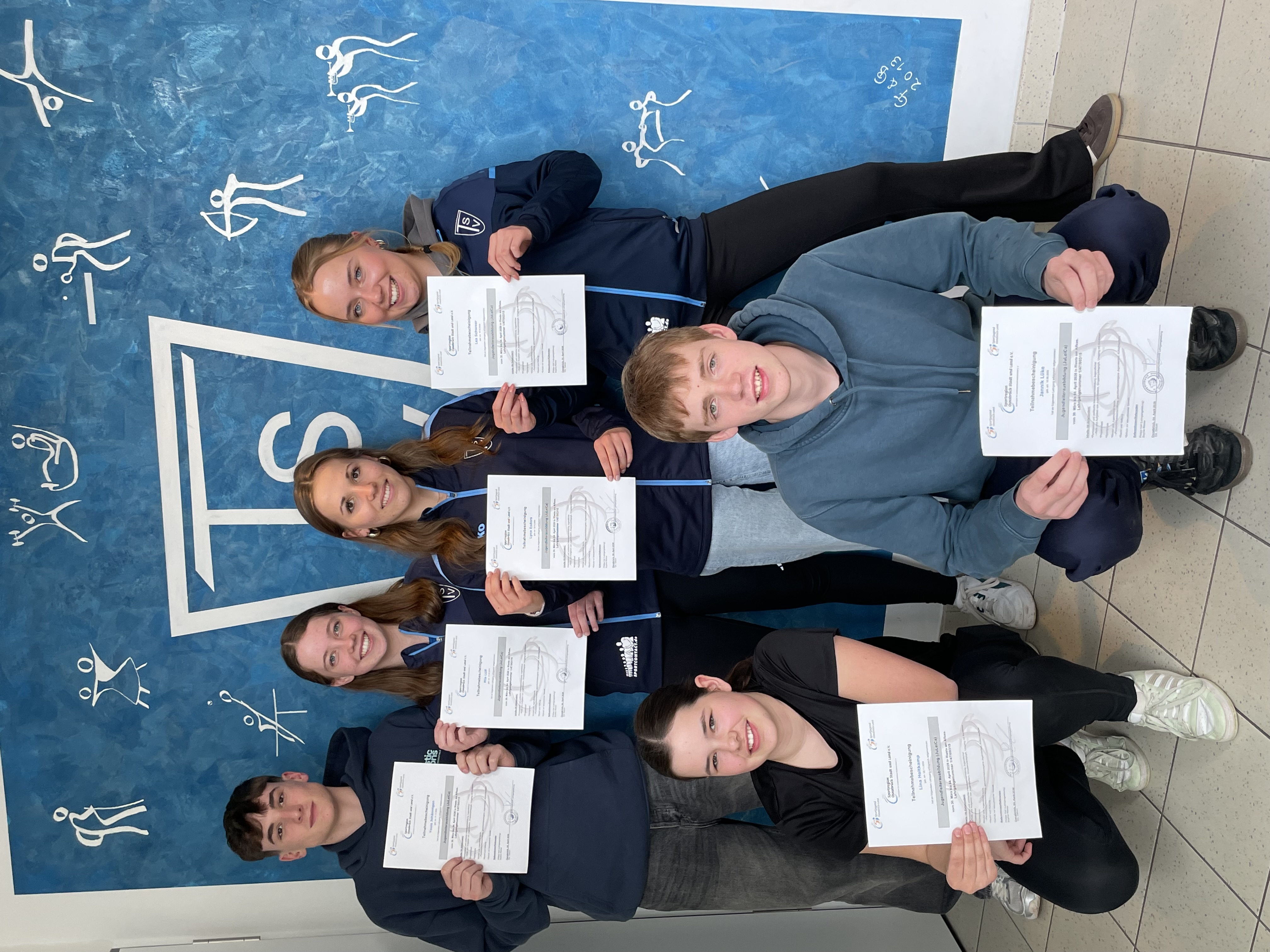 6 neue Juleica Inhaber für den TSV Westerhausen Auf dem Foto sind sechs Jugendliche zu sehen, die gemeinsam für ein Gruppenbild posieren 📸. Vier stehen in der hinteren Reihe, zwei knien vorne. Alle halten jeweils eine Urkunde in den Händen und schauen in die Kamera. Die Urkunden wirken wie Teilnahme- oder Qualifikationsbescheinigungen. Die Jugendlichen tragen überwiegend sportliche Kleidung. Drei Personen in der hinteren Reihe tragen dunkelblaue Trainingsjacken mit Vereinslogo, was auf eine Zugehörigkeit zu einem Sportverein hindeutet. Die beiden Personen vorne tragen Freizeit- bzw. Sportkleidung (Hoodie bzw. T-Shirt). Im Hintergrund befindet sich eine große blaue Wand mit einem stilisierten Vereinslogo („TSV“) in der Mitte. Rundherum sind weiße Piktogramme verschiedener Sportarten dargestellt, zum Beispiel Turnen, Leichtathletik, Bogenschießen und Wintersportarten 🏃♀️🏹⛷️. Der Hintergrund vermittelt deutlich den Eindruck einer Sportvereins- oder Ausbildungsumgebung. Die Gruppe wirkt zufrieden und stolz auf ihre Urkunden, was darauf schließen lässt, dass sie erfolgreich an einer Schulung oder Qualifikation teilgenommen haben 🎓.