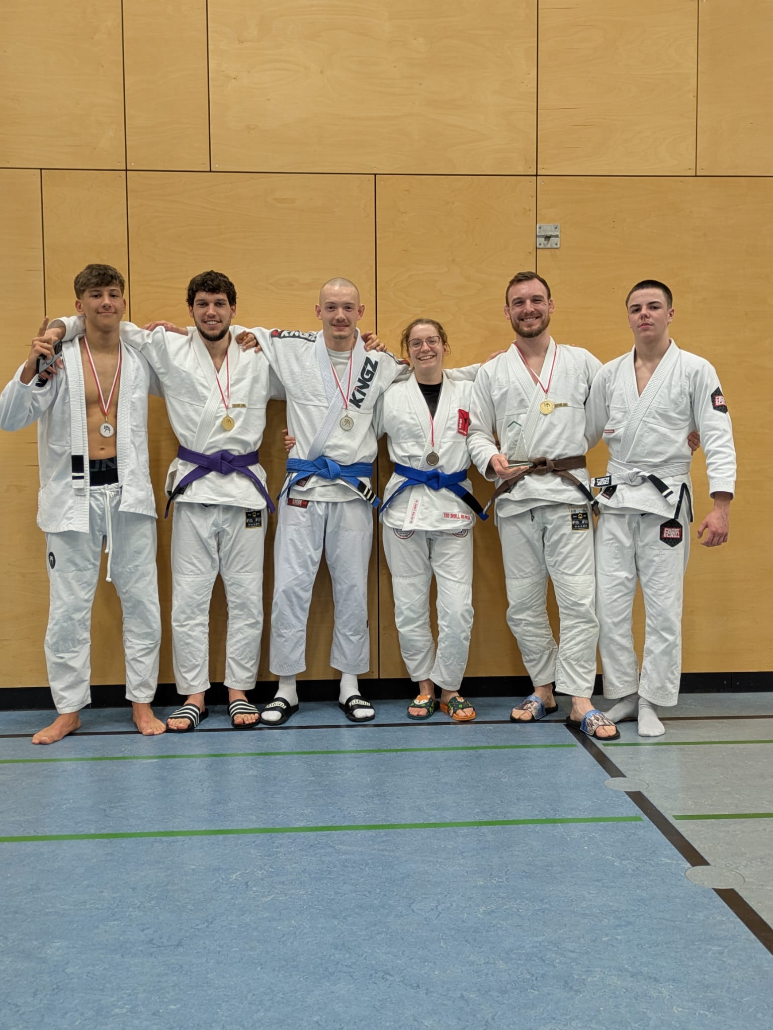 TSV Kampfsportler beim Lippe Cup in Lemgo am 31.01.2026 Das Bild zeigt ein Gruppenfoto von sechs jungen Erwachsenen nach einem Kampfsport-Wettkampf, vermutlich Judo oder Brazilian Jiu-Jitsu. Alle Personen stehen nebeneinander vor einer hellen Holzpaneelwand in einer Sporthalle. Sie tragen weiße Kampfanzüge (Gi) mit unterschiedlich farbigen Gürteln (u. a. weiß, blau, lila und braun), was auf verschiedene Leistungs- oder Erfahrungsstufen hinweist. Mehrere Personen tragen Medaillen um den Hals, eine Person hält zusätzlich eine Urkunde oder Auszeichnung in der Hand. Die Gruppe wirkt stolz, zufrieden und kameradschaftlich – einige lächeln, andere schauen ruhig in die Kamera, viele haben den Arm um die Schulter der Nachbarperson gelegt. Auffällig ist die entspannte Haltung: Einige tragen Badelatschen, andere stehen barfuß, was typisch für Kampfsportveranstaltungen ist. Der Hallenboden ist blau-grau mit grünen Linien, im Hintergrund dominiert die schlichte Holzverkleidung der Wand. Insgesamt vermittelt das Foto den Eindruck eines erfolgreichen Wettkampftages, Teamzusammenhalt und sportlicher Fairness.