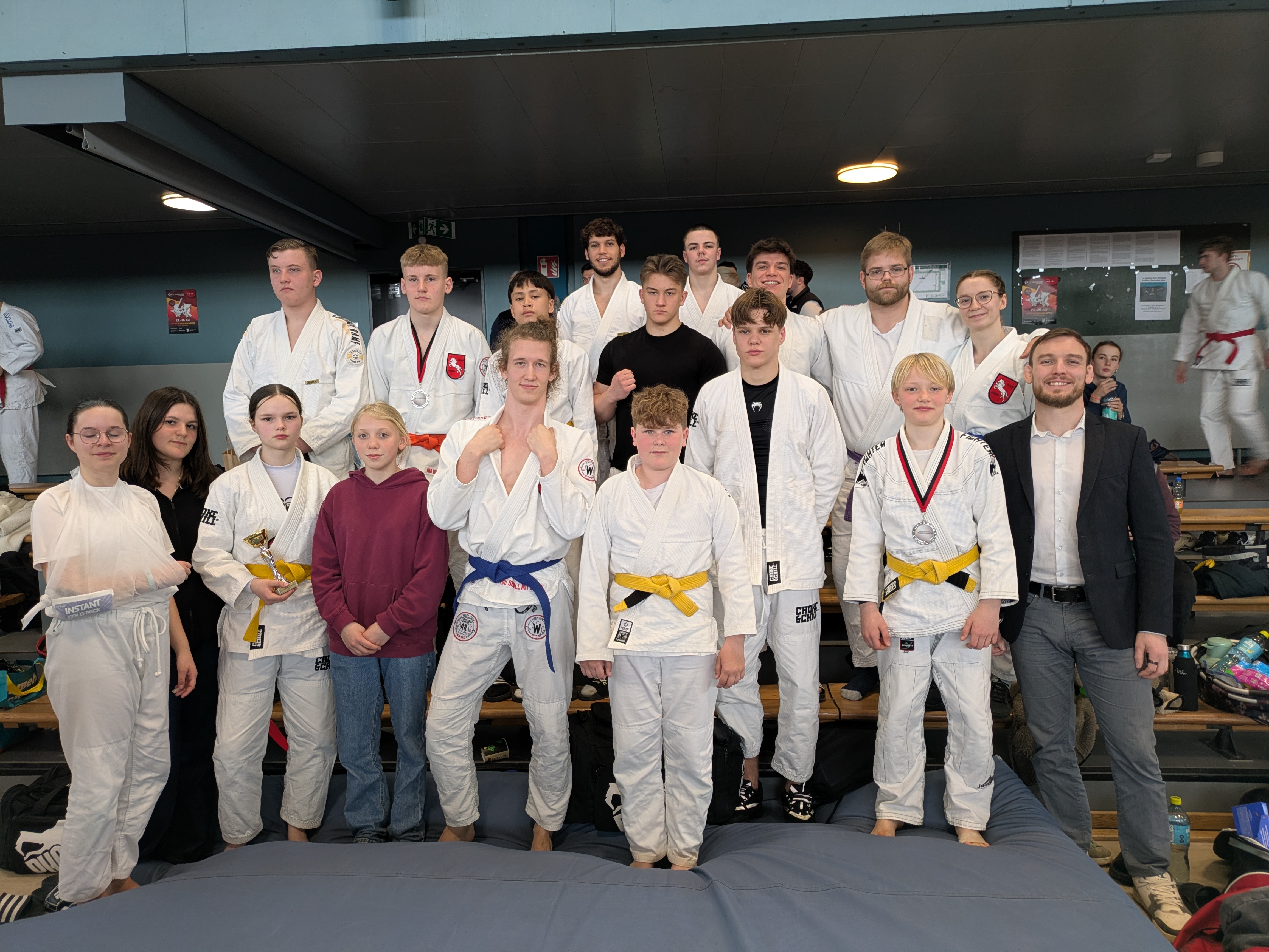 Norddeutsche Meisterschaft JuJutsu in Hamburg 17. + 18.04.2026 Gruppe von Jugendlichen und jungen Erwachsenen in Judoanzügen mit gelben und blauen Gürteln sowie in Freizeitkleidung vor einer Wand in einer Sporthalle