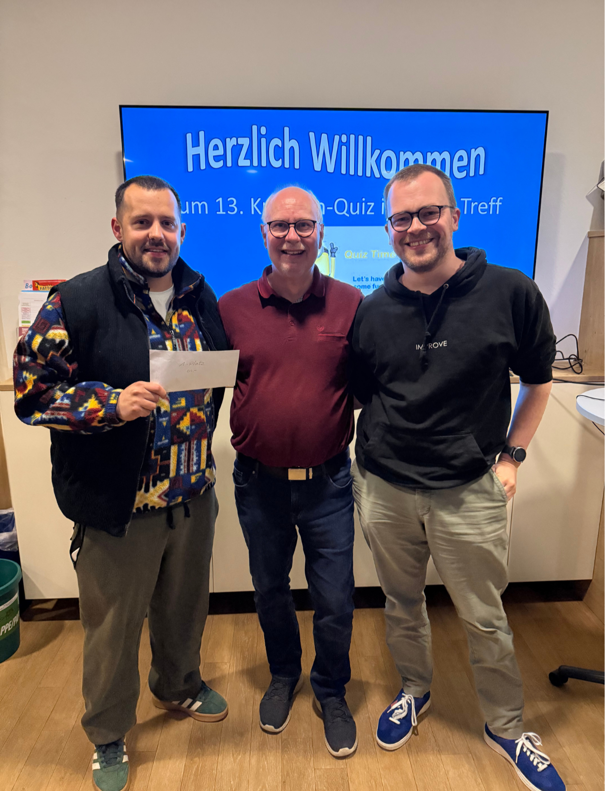 Sieger Kneipenquiz 20.03.2026 Das Bild zeigt drei Männer, die nebeneinander stehen und in die Kamera lächeln. Sie befinden sich offenbar in einem Innenraum, möglicherweise bei einer Veranstaltung oder einem Treffen. Hinter ihnen ist ein großer Bildschirm zu sehen mit der Aufschrift „Herzlich Willkommen“ sowie weiteren deutschen Texten, die auf ein Quiz-Event oder Treffen hinweisen (z. B. „Quiz Time“). Der Mann links trägt eine gemusterte, bunte Jacke unter einer dunklen Weste und hält einen Umschlag in der Hand – möglicherweise ein Preis oder eine Auszeichnung. Der Mann in der Mitte trägt ein rotes Poloshirt und eine Brille. Der Mann rechts trägt einen schwarzen Hoodie, eine Brille und helle Hosen. Alle drei wirken entspannt und gut gelaunt, als ob sie gerade an einem gemeinsamen Event teilnehmen oder etwas gewonnen haben.
