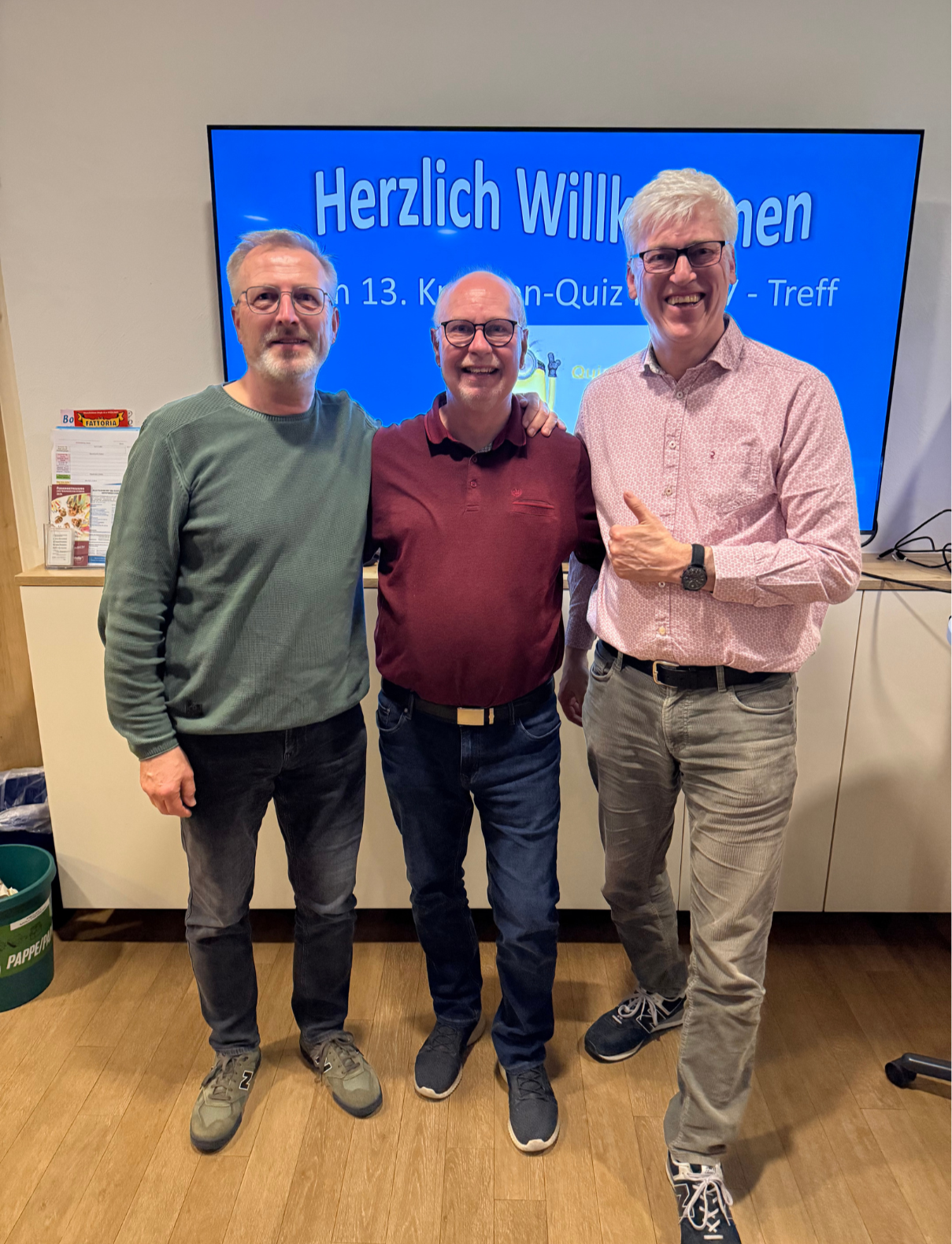 2. Platz Kneipenquiz 20.03.2026 Das Bild zeigt drei Männer, die nebeneinander stehen und in die Kamera lächeln. Sie wirken entspannt und freundlich, vermutlich bei einer gemeinsamen Veranstaltung. Im Hintergrund ist ein großer Bildschirm mit blauem Hintergrund zu sehen. Darauf steht auf Deutsch „Herzlich Willkommen“ sowie Hinweise auf ein „Quiz“ und ein „Treff“, was darauf hindeutet, dass es sich um ein Quiz-Event oder ein geselliges Treffen handelt. Der Mann links trägt einen grünen Pullover, Jeans und Sneaker. Er hat kurze graue Haare, einen Bart und eine Brille. Der Mann in der Mitte trägt ein bordeauxrotes Polo-Shirt, Jeans und dunkle Schuhe. Er ist ebenfalls Brillenträger und lächelt breit. Der Mann rechts trägt ein helles, gemustertes Hemd, graue Jeans und Sneaker. Er hat hellblondes Haar, eine Brille und zeigt mit der Hand eine freundliche Geste (Daumen hoch). Alle drei stehen nah beieinander, zwei haben die Arme locker umeinander gelegt, was auf eine vertraute oder kollegiale Beziehung schließen lässt. Die Umgebung wirkt wie ein moderner Innenraum, möglicherweise ein Büro, ein Vereinsraum oder ein Veranstaltungsraum.