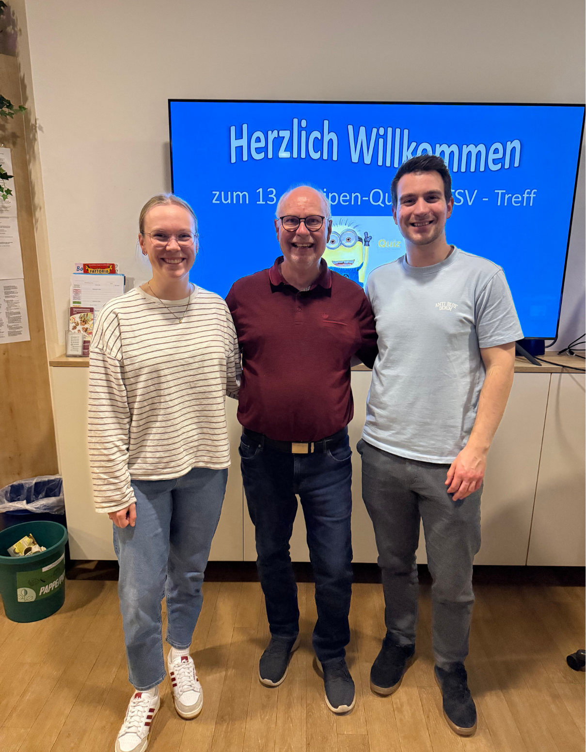 Kneipenquiz 20.03.2026 3. Platz Das Bild zeigt drei Personen, die nebeneinander stehen und freundlich in die Kamera lächeln. Sie befinden sich in einem Innenraum, vermutlich bei einer Veranstaltung oder einem Treffen. Im Hintergrund ist ein großer Bildschirm mit blauem Hintergrund zu sehen. Darauf steht deutlich „Herzlich Willkommen“ sowie weiterer deutscher Text, der auf ein „Kneipen-Quiz“ oder ein Treffen hinweist. Neben dem Text ist eine kleine Comicfigur zu erkennen, die zum Quiz-Thema passt. Links steht eine Frau mit zusammengebundenen Haaren und Brille. Sie trägt ein helles, gestreiftes Langarmshirt, eine blaue Jeans und weiße Sneaker. In der Mitte steht ein älterer Mann mit Brille, der ein rotes Poloshirt, eine dunkle Jeans und dunkle Schuhe trägt. Rechts steht ein jüngerer Mann mit kurzem dunklem Haar. Er trägt ein hellblaues T-Shirt, eine graue Hose und dunkle Schuhe. Alle drei wirken entspannt und gut gelaunt, als ob sie gemeinsam an dem Event teilnehmen oder gerade ein Foto zur Erinnerung machen.