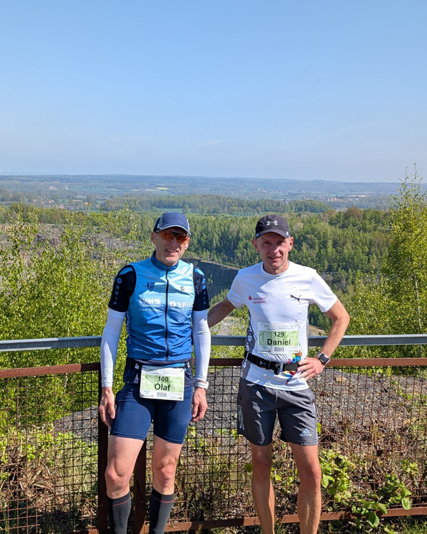 14. Osnabrücker Gifpelsturm 1 Zwei Personen stehen nebeneinander an einem Metallgeländer mit Blick auf eine weite, grüne Landschaft. Beide tragen Sportbekleidung, Startnummern und Sonnenbrillen. Die linke Person trägt ein blaues Trikot und eine kurze Hose, die rechte Person ein weißes T-Shirt und eine graue kurze Hose. Im Hintergrund sind Bäume und ein weiter Horizont zu sehen.