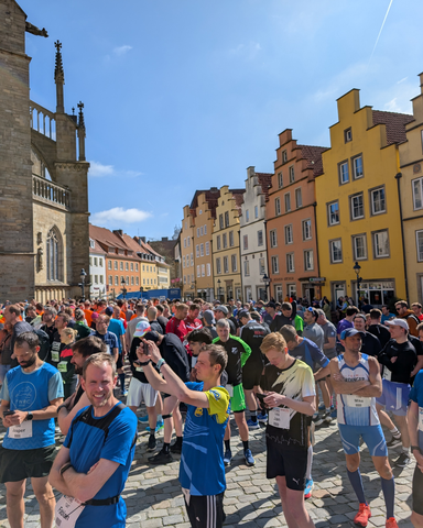 14. Osnabrücker Gipfelsturm 2 Eine große Menschenmenge steht auf einem gepflasterten Platz vor einer Reihe farbenfroher, giebelständiger Häuser. Viele Personen tragen Sportkleidung und Startnummern, einige fotografieren mit dem Smartphone. Im linken Bildbereich ist ein Teil eines historischen Gebäudes mit gotischen Fenstern zu sehen. Die Szene spielt bei sonnigem Wetter unter blauem Himmel.
