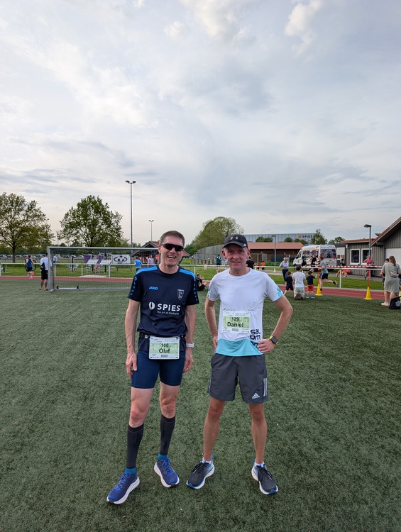 Bifurkationslauf Gesmold 1 Zwei Personen stehen auf einem Sportplatz mit Kunstrasen. Beide tragen Sportkleidung und Startnummern an der Brust. Im Hintergrund sind weitere Menschen, ein Fußballtor, Absperrbänder und ein Gebäude zu sehen. Der Himmel ist bewölkt.