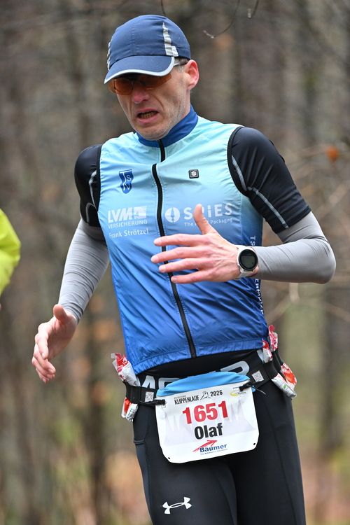 Olaf Meyer beim Klippenlauf Das Bild zeigt eine Nahaufnahme eines Läufers während eines Wettkampfs im Wald. Der Mann befindet sich in Bewegung und wirkt deutlich angestrengt: Sein Gesicht ist angespannt, der Mund leicht geöffnet, als würde er schwer atmen. Er trägt eine blaue Kappe mit hellem Streifen sowie eine orange getönte Sportbrille. Sein Oberkörper ist mit einer blau verlaufenden, ärmellosen Laufweste bekleidet, unter der er ein schwarzes Kurzarmshirt und graue Armstulpen trägt. Auf der Weste sind Logos und Schriftzüge zu erkennen. An seinem linken Handgelenk trägt er eine Sportuhr. Um die Hüfte hat er einen Laufgürtel mit mehreren Energie-Gels verstaut, die seitlich sichtbar sind. An der Vorderseite ist seine Startnummer befestigt: „1651“ mit dem Namen „Olaf“. Dazu trägt er schwarze, enganliegende Laufhosen. Der Hintergrund ist unscharf und zeigt einen herbstlichen Wald mit kahlen Bäumen und gedämpften Farben. Die Fokussierung auf den Läufer verstärkt den Eindruck von Intensität und Ausdauer während eines anspruchsvollen Geländelaufs.