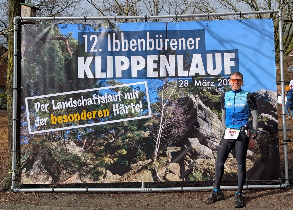 12. Ibbenbürener Klippenlauf Das Bild zeigt ein großes Veranstaltungsbanner im Freien, das an einem Metallrahmen befestigt ist. Im Hintergrund erkennt man Bäume ohne Blätter, was auf eine kühle Jahreszeit hindeutet. Auf dem Banner steht in großen, auffälligen Buchstaben: „12. Ibbenbürener KLIPPENLAUF“ sowie das Datum „28. März 20..“ (die letzte Jahreszahl ist nicht vollständig sichtbar). Darunter ist der Slogan zu lesen: „Der Landschaftslauf mit der besonderen Härte!“, wobei das Wort „besonderen“ farblich hervorgehoben ist. Das Banner zeigt außerdem eine Landschaft mit Felsen, Bäumen und einem Waldgebiet, was auf einen anspruchsvollen Lauf durch unebenes Gelände hinweist. Rechts vor dem Banner steht ein Mann in Sportkleidung. Er trägt eine enge Laufhose, ein langärmliges Oberteil mit Weste sowie eine Startnummer mit der Zahl 1651 und dem Namen „Olaf“. Er hat eine Sonnenbrille auf, lächelt und wirkt zufrieden oder stolz, vermutlich nach der Teilnahme am Lauf. Seine Haltung ist entspannt, mit leicht angewinkelten Armen. Insgesamt vermittelt das Bild die Atmosphäre eines sportlichen Outdoor-Events in einer natürlichen, etwas rauen Umgebung.