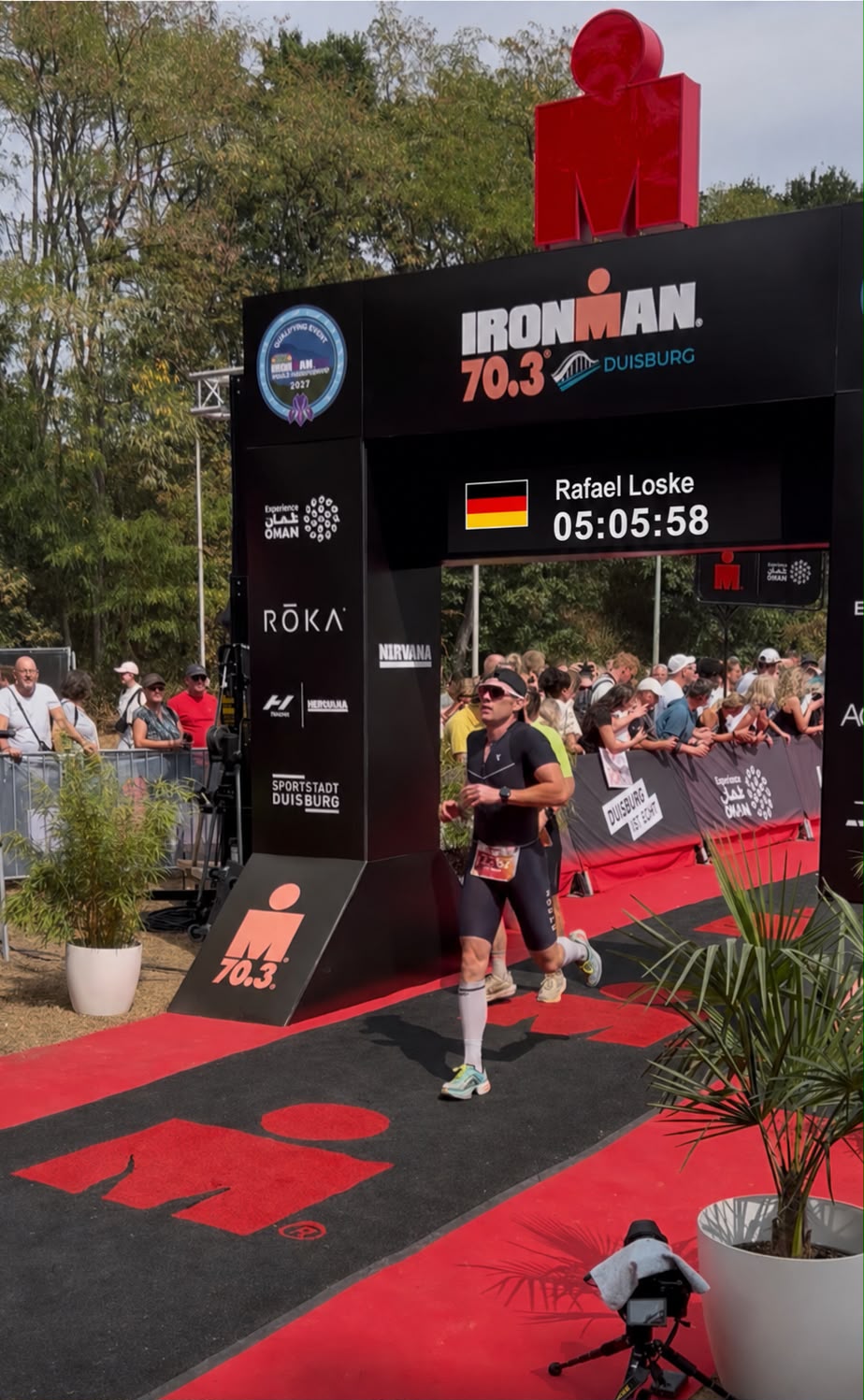 Zieleinlauf Iron Man 16.08.2026 Athlet läuft auf rotem Teppich unter schwarzem Zielbogen mit rotem Ironman-Logo und digitaler Zeitnahme bei Ironman 70.3 Duisburg