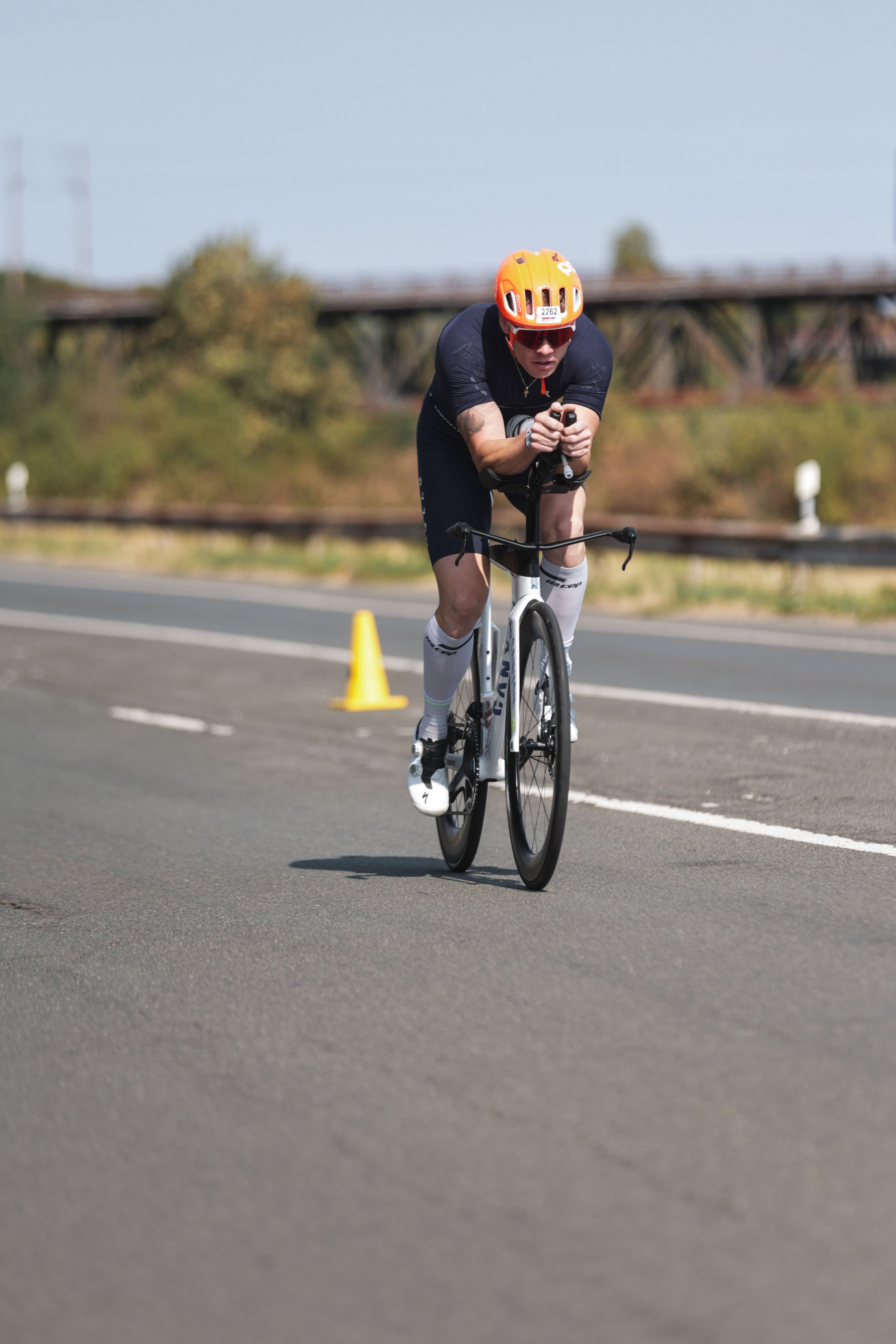 Disziplin Radfahren - Iron Man 16.08.2026 Radrennfahrer mit orangefarbenem Helm in aerodynamischer Haltung auf Rennrad auf asphaltierter Straße mit gelbem Verkehrshütchen im Hintergrund