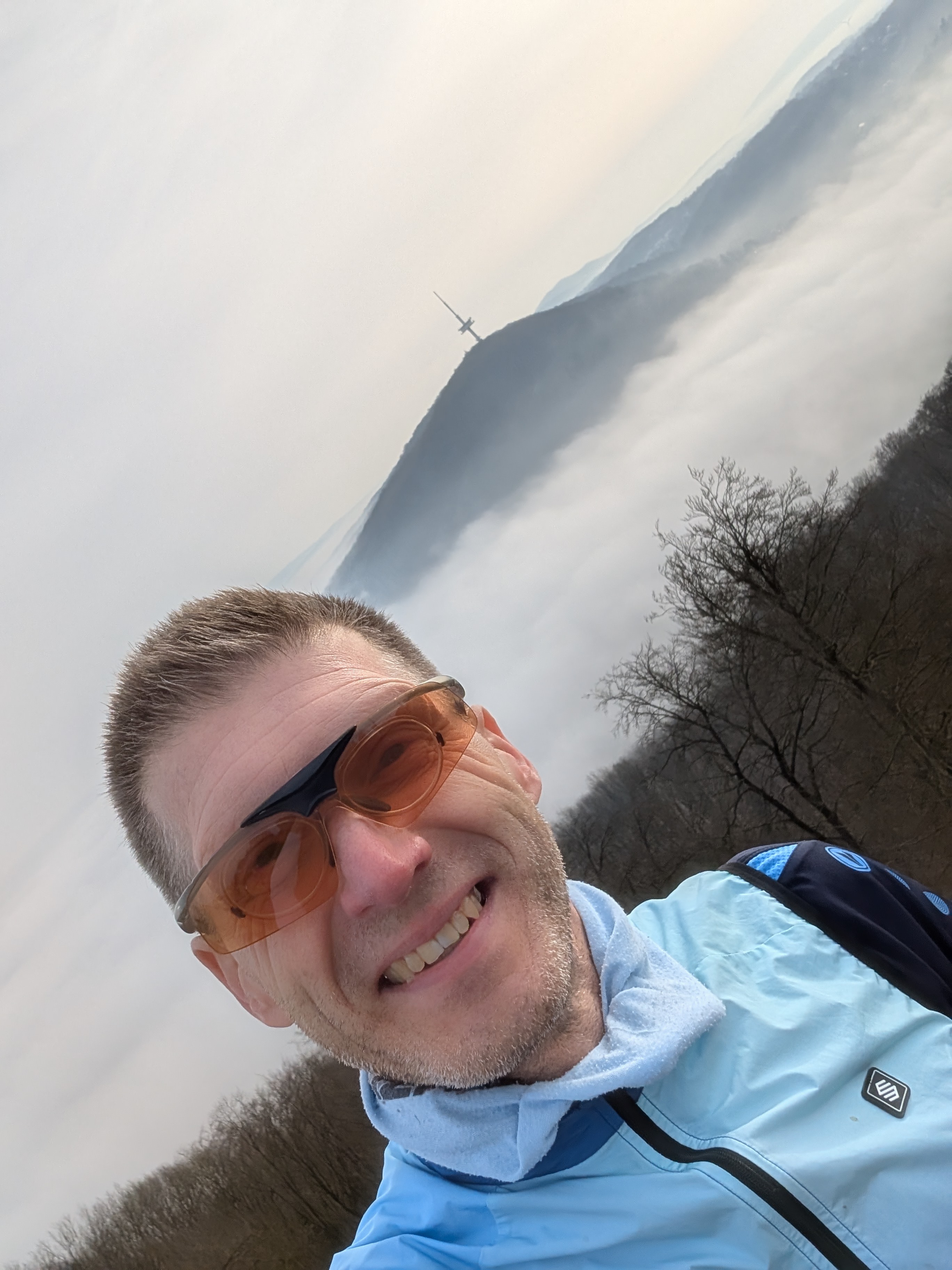Wittekindslauf - Olaf Meyer Das Bild zeigt ein Selfie eines Mannes in einer bergigen Landschaft. ⛰️ Vordergrund: Ein Mann mit kurzen Haaren und leichtem Bart lächelt in die Kamera. Er trägt eine Sportjacke in Hellblau, eine Sonnenbrille mit orangefarbenen Gläsern und hat einen Rucksack auf dem Rücken. Sein Gesicht wirkt entspannt und zufrieden. Hintergrund: Im Hintergrund erstreckt sich eine Hügellandschaft oder ein Mittelgebirge, das teilweise von Nebel oder tiefhängenden Wolken bedeckt ist. Auf einem Bergrücken ist ein großes Kreuz (Gipfelkreuz) zu erkennen. Außerdem sind kahle Bäume zu sehen, was auf eine kühlere Jahreszeit hindeutet. Besonderheit: Das Bild ist leicht schräg aufgenommen, was dem Foto eine dynamische Perspektive verleiht. Gesamteindruck: Die Szene vermittelt den Eindruck einer Wanderung oder eines Outdoor-Ausflugs in ruhiger Natur, mit einer schönen Aussicht über eine nebelverhangene Landschaft.