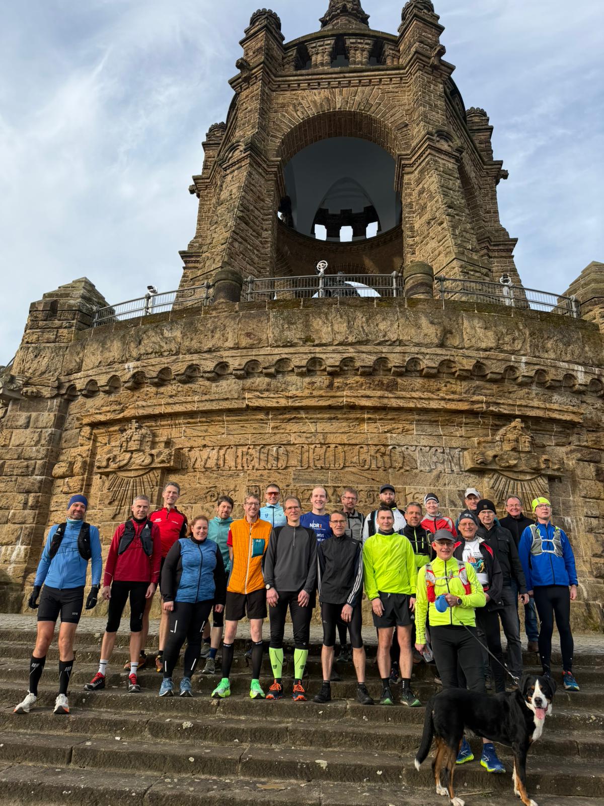 Start Wittekindslauf Das Bild zeigt eine Gruppe von etwa 20 Personen, die gemeinsam vor einem großen, historischen Bauwerk posieren. Es handelt sich um ein monumentales steinernes Aussichtsbauwerk mit einem turmartigen Aufbau und einem großen Rundbogen in der Mitte. Die Architektur wirkt massiv und erinnert an ein Denkmal oder einen Aussichtsturm. Die Gruppe steht auf breiten Steinstufen direkt vor einer Mauer, in die der Schriftzug „Wilhelm der Großen“ (teilweise sichtbar) eingraviert ist. Viele der Personen tragen Sportkleidung wie Laufjacken, Tights, Shorts und Laufschuhe, was darauf hindeutet, dass es sich um eine Lauf- oder Wandergruppe handelt. Die Kleidung ist bunt und funktional, passend zu kühlerem Wetter. Die Menschen wirken entspannt und gut gelaunt, einige lächeln in die Kamera. Vorne rechts steht eine Person in auffälliger neongelber Jacke, daneben ein schwarzer Hund mit weißer Brust, der ebenfalls in Richtung Kamera schaut. Im Hintergrund ragt das Bauwerk imposant nach oben, mit einem Geländer und Aussichtsplattform sowie dekorativen Steinverzierungen. Der Himmel ist leicht bewölkt, aber hell, was dem Bild eine klare und freundliche Stimmung verleiht.