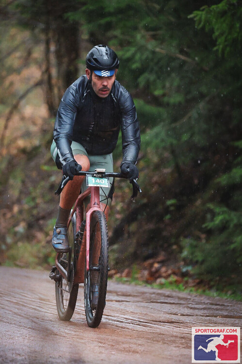 Harzgravel 2 Ein Radfahrer mit dunkler, enganliegender Regenjacke, kurzer Hose und Helm fährt auf einem nassen, unbefestigten Waldweg. Die Umgebung ist von dichtem, grünem Nadelwald geprägt. Am unteren Bildrand befindet sich ein farbiges Logo mit einer stilisierten Landkarte und dem Schriftzug 'SPORTOGRAF.COM'.