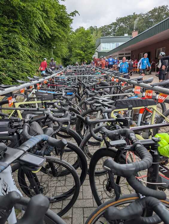 Beast of Bramsche 1 Dicht an dicht abgestellte Rennräder stehen in mehreren Reihen auf einem gepflasterten Bereich. Jedes Fahrrad ist mit einer Startnummer am Lenker versehen. Im Hintergrund sind zahlreiche Menschen in Sportkleidung zu sehen, einige tragen blaue Oberteile. Links begrenzt eine dichte Baumreihe das Bild, rechts steht ein modernes Gebäude mit großen Fenstern und einer Überdachung.