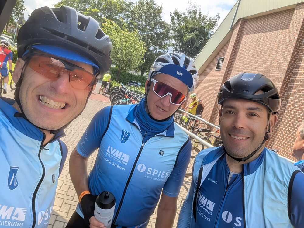 Beast of Bramsche 2 Drei Personen in hellblauen Radtrikots und dunklen Fahrradhelmen stehen nebeneinander. Im Hintergrund sind weitere Menschen, Fahrräder und ein Gebäude mit roter Klinkerfassade zu sehen. Eine der Personen hält eine weiße Trinkflasche mit schwarzem Deckel in der Hand. Die Trikots zeigen verschiedene Logos und Schriftzüge.