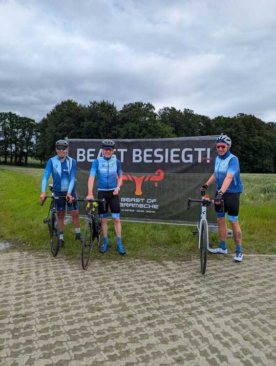 Beast of Bramsche 3 Drei Personen in hellblauen Radtrikots und dunklen Fahrradhelmen stehen nebeneinander. Im Hintergrund sind weitere Menschen, Fahrräder und ein Gebäude mit roter Klinkerfassade zu sehen. Eine der Personen hält eine weiße Trinkflasche mit schwarzem Deckel in der Hand. Die Trikots zeigen verschiedene Logos und Schriftzüge.