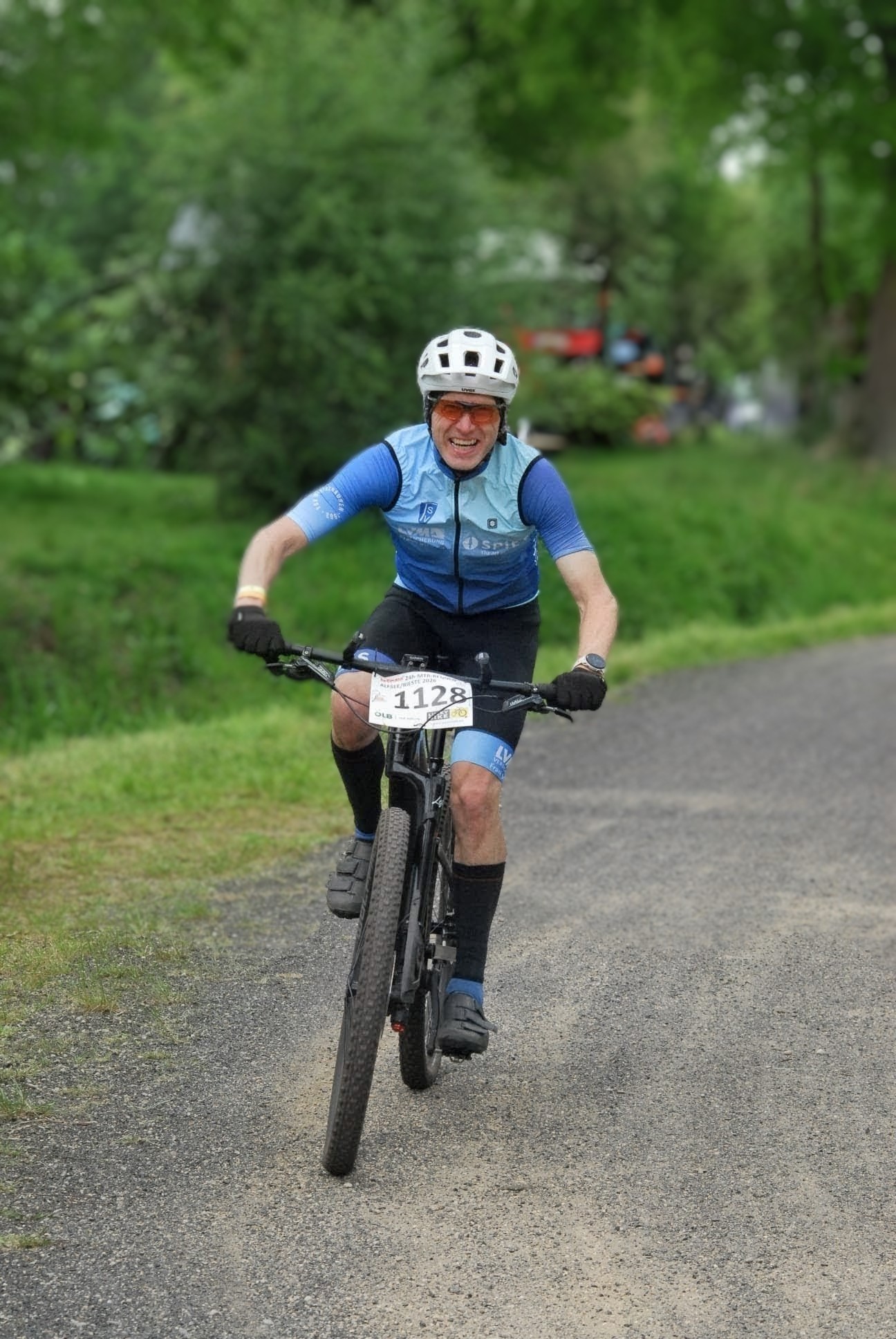 Olaf Meyer Das Bild zeigt eine Person bei einem Mountainbike-Rennen auf einem Schotterweg in einer grünen, parkähnlichen Umgebung. Die Person trägt einen weißen Fahrradhelm, eine Sportbrille mit orangefarbenen Gläsern sowie ein blaues Radtrikot und schwarze Handschuhe. Auf dem Lenker ist eine Startnummer mit der Nummer „1128“ befestigt. Die Person fährt dynamisch auf die Kamera zu und lächelt bzw. wirkt angestrengt, aber motiviert. Der Hintergrund ist unscharf, wodurch der Fokus klar auf dem Radfahrer liegt. Die Szene vermittelt Bewegung, Sportlichkeit und Freude am Radfahren in der Natur.