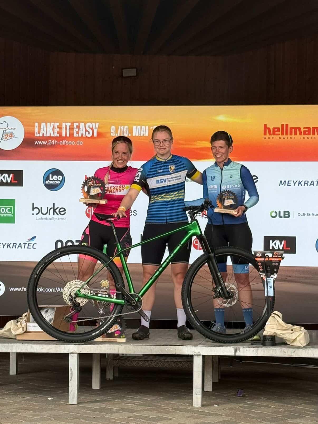 Siegerehrung Frauen Das Bild zeigt eine Siegerehrung bei einem Mountainbike- oder Radsportevent. Drei Personen stehen auf einer Bühne vor einer großen Sponsorenwand mit dem Schriftzug „LAKE IT EASY“ und dem Datum „9./10. Mai“. Die Person links trägt ein auffälliges pink-schwarzes Radtrikot und hält einen Pokal. In der Mitte steht eine Person im blau-schwarzen Vereinsdress neben einem grünen Mountainbike. Rechts steht eine weitere Person in einem blau-hellblauen Radtrikot und hält ebenfalls einen Pokal. Alle drei lächeln in die Kamera. Vor ihnen steht das grüne Fahrrad prominent auf der Bühne, was darauf hindeutet, dass es sich möglicherweise um einen Gewinn oder ein besonderes Präsent handelt. Die Stimmung wirkt feierlich und sportlich, typisch für eine Preisverleihung nach einem Wettkampf.