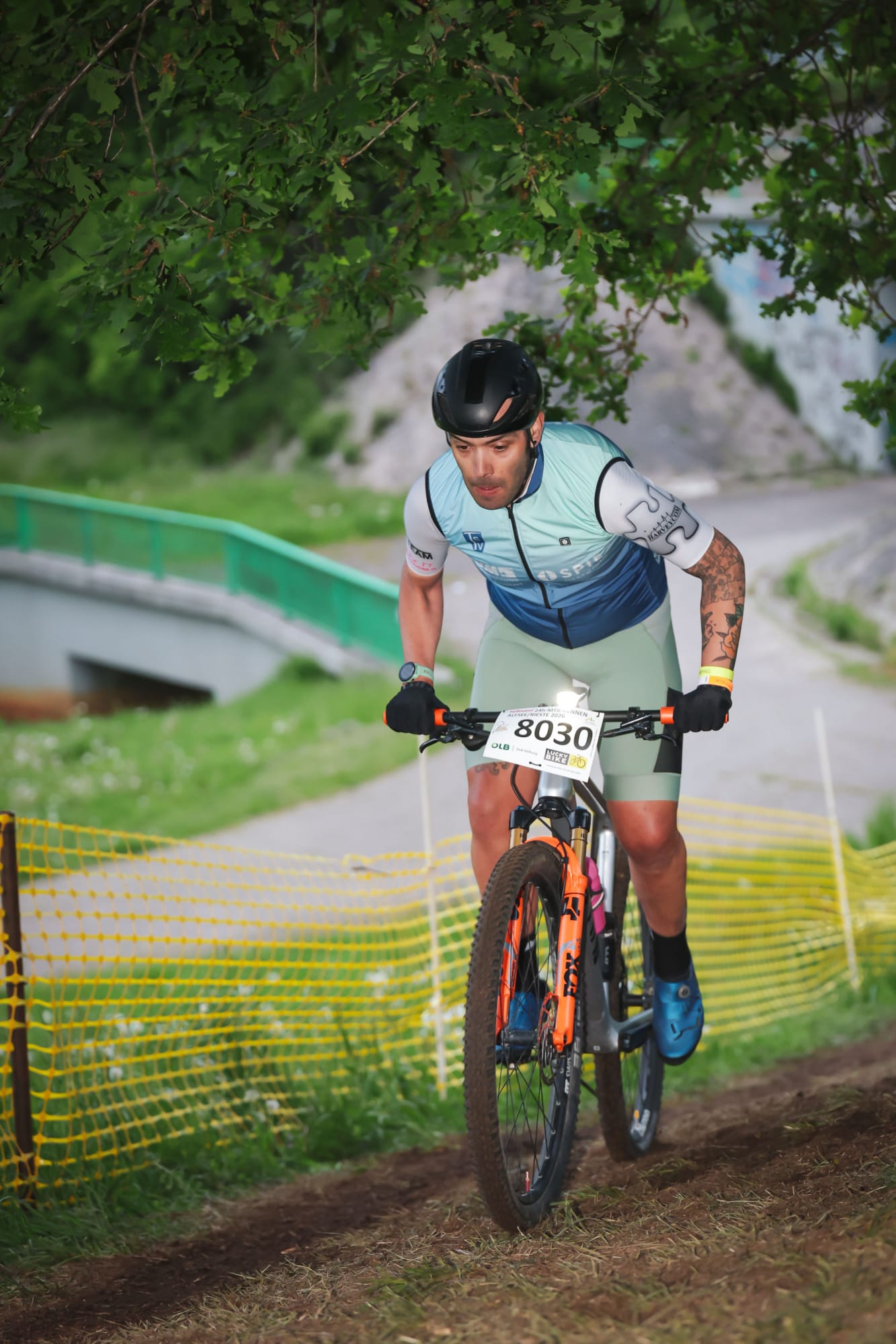 Das Bild zeigt einen Mountainbiker während eines Rennens auf einem schmalen Erdtrail in einer grünen Naturkulisse. Die Person trägt einen schwarzen Fahrradhelm, ein blau-weißes Radtrikot, kurze helle Radhosen sowie schwarze Handschuhe. Auffällig sind die Tätowierungen am rechten Arm und die blauen Fahrradschuhe. Auf dem Lenker ist die Startnummer „8030“ befestigt.  Der Fahrer wirkt konzentriert und kraftvoll, während er leicht bergauf fährt. Das Mountainbike besitzt eine auffällige orangefarbene Federgabel und breite Geländereifen. Entlang der Strecke verläuft ein gelbes Absperrnetz. Im Hintergrund sind eine kleine Brücke, ein Weg und dichter grüner Bewuchs zu erkennen. Die überhängenden Äste rahmen die Szene ein und verstärken die natürliche Atmosphäre.  Durch die unscharfe Hintergrunddarstellung hebt sich der Fahrer deutlich hervor, wodurch das Bild Dynamik, Geschwindigkeit und Wettkampfcharakter vermittelt.