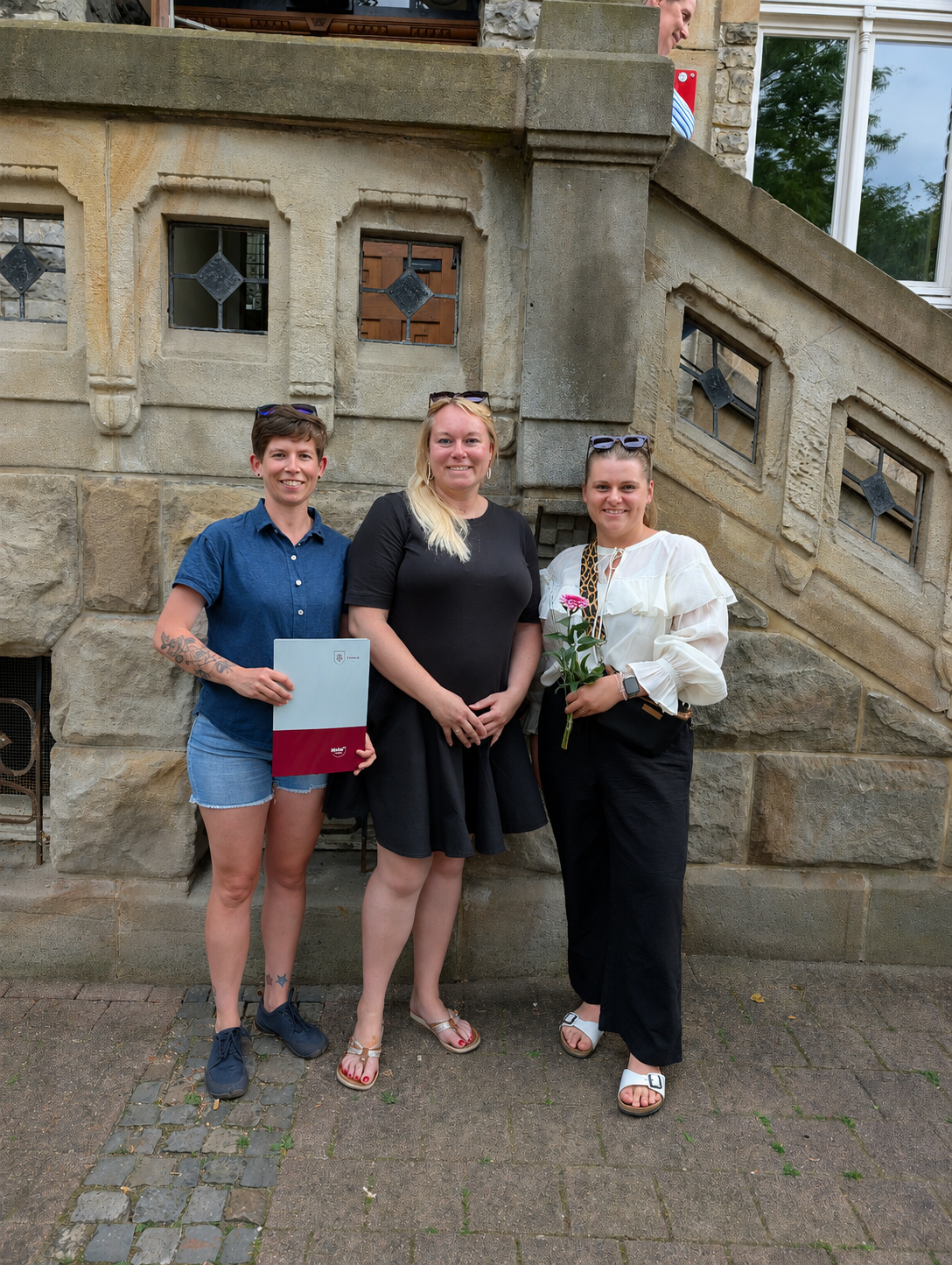 Prämierung Stadtradeln 2026 Drei Frauen stehen vor einer steinernen Treppe mit verzierten Fenstern, eine hält eine Urkunde, eine andere eine Rose