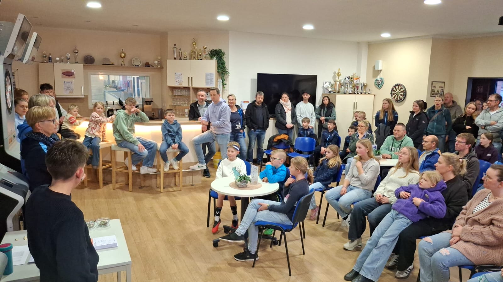 Verleihung Sportabzeichen 03.03.2026 - Bild 3 In einem hellen Vereinsraum sitzen und stehen viele Kinder, Jugendliche und Erwachsene und hören aufmerksam einer Person zu, die vorne spricht. Einige Kinder sitzen auf Barhockern an einer Theke, andere auf Stühlen an kleinen Tischen. An den Wänden hängen Dartscheiben, und auf Schränken stehen zahlreiche Pokale. Die Atmosphäre wirkt wie eine gemeinsame Vereinsveranstaltung oder Ehrung.