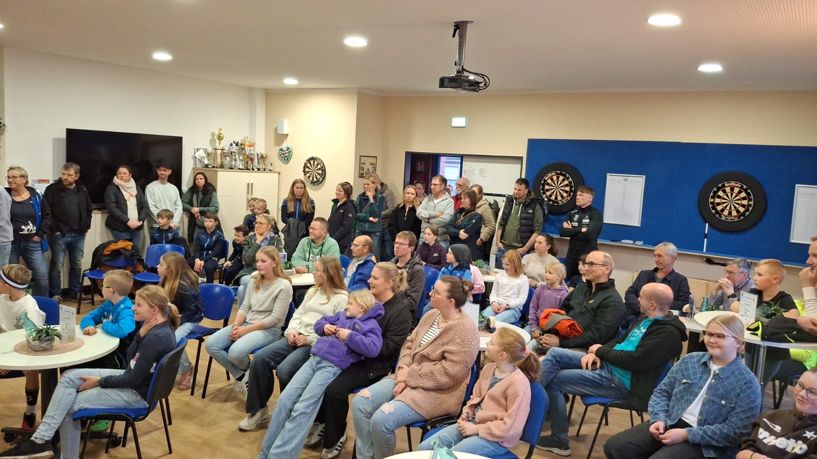Verleihung Sportabzeichen 03.03.2026 - Bild 2 Ein Raum mit vielen Kindern, Jugendlichen und Erwachsenen, die auf Stühlen und an Tischen sitzen oder stehen. Sie schauen nach vorne und hören aufmerksam zu. An der Wand hängen zwei Dartscheiben, daneben stehen Pokale auf einem Schrank.