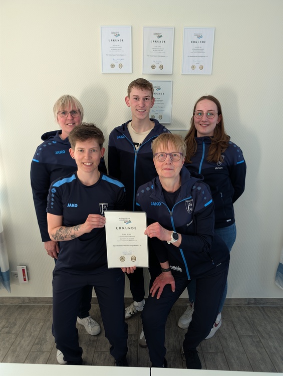 Sportabzeichenwettbewerb Sechs Personen in einheitlicher, dunkelblauer Sportkleidung stehen und sitzen in einem Raum mit hellen Wänden. Die vordere Person hält eine Urkunde mit dem Titel 'URKUNDE' in der Hand. Im Hintergrund hängen vier weitere Urkunden gerahmt an der Wand. Die Sportkleidung trägt das Logo und den Schriftzug eines Sportvereins auf der Brust sowie den Markennamen 'JAKO' auf der rechten Schulter. Der Boden ist mit grauen Fliesen ausgelegt.