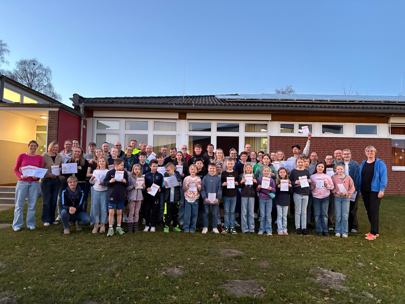 Gruppenfoto Verleihung Sportabzeichen am 03.03.2026 Großes Gruppenfoto vor einem Vereinsgebäude: Viele Kinder, Jugendliche und Erwachsene stehen gemeinsam auf einer Wiese und halten Urkunden in den Händen. Die Gruppe blickt in die Kamera und wirkt fröhlich und stolz. Einige Personen halten ihre Urkunden hoch. Im Hintergrund ist das Gebäude mit Fenstern und Solarpanelen auf dem Dach zu sehen.