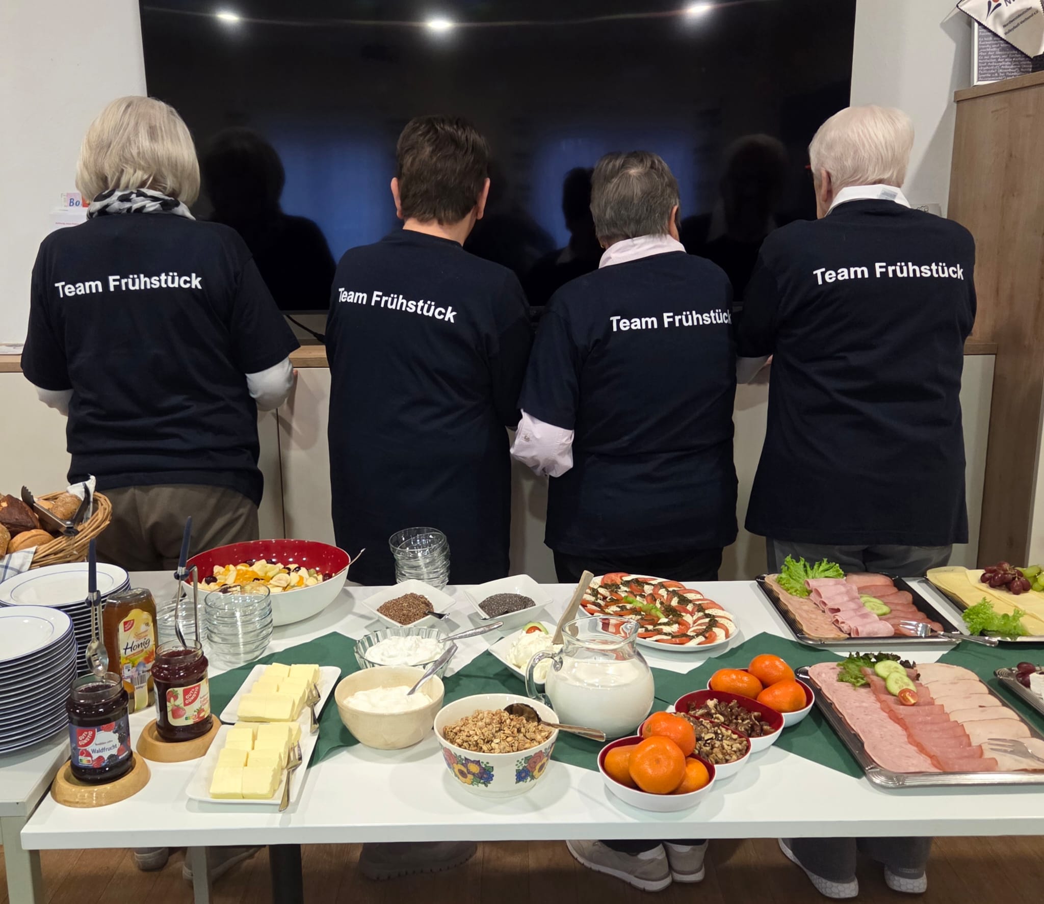 Fit und Frühstück Vier Personen mit schwarzen T-Shirts mit der Aufschrift 'Team Frühstück' stehen mit dem Rücken zu einem großen Bildschirm vor einem Tisch mit verschiedenen Frühstückszutaten und -gerichten
