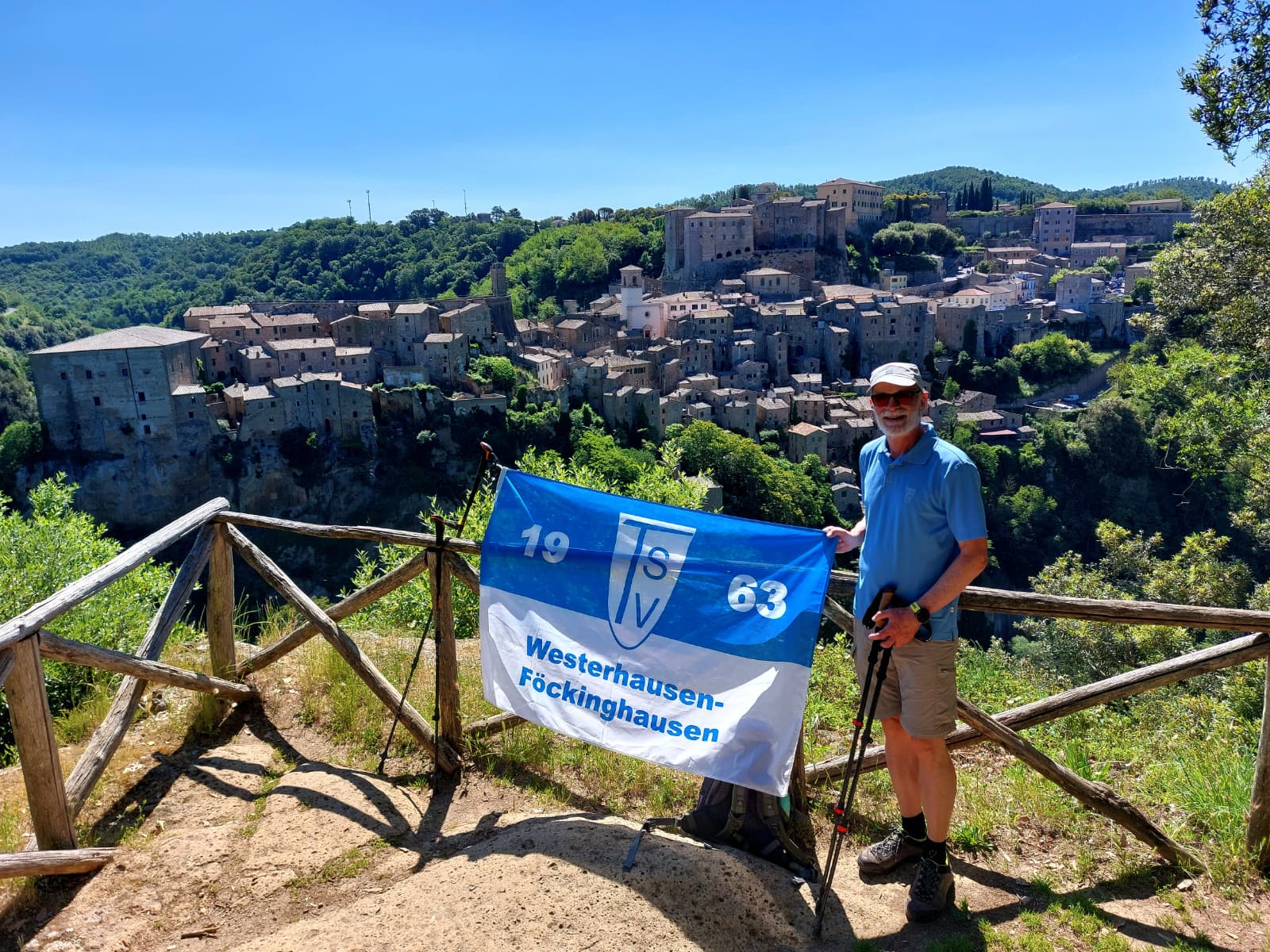 TSV on Tour - Grüße aus der Toskana Das Bild zeigt einen Wanderer auf einem Aussichtspunkt mit Blick auf eine historische italienische Bergstadt in der Toskana. Der Mann trägt ein blaues Poloshirt, kurze Wanderhose, Wanderschuhe, Sonnenbrille und eine helle Kappe. In den Händen hält er Wanderstöcke. Vor ihm ist eine blau-weiße Fahne mit der Aufschrift „SV 1963 Westerhausen-Föckinghausen“ befestigt. Im Hintergrund erstreckt sich eine malerische mittelalterliche Stadt aus Natursteinhäusern auf einem Felsen, umgeben von grünen Hügeln und dichter Vegetation. Besonders auffällig sind die alten Gebäude, die Kirchenkuppel und die befestigten Mauern auf der Anhöhe. Das Wetter ist sonnig, der Himmel wolkenlos blau, wodurch die Landschaft sehr freundlich und sommerlich wirkt. Die Aufnahme vermittelt den Eindruck einer Wanderreise oder eines Ausflugs in die Toskana, vermutlich bei Sorano oder einer ähnlichen historischen Ortschaft.
