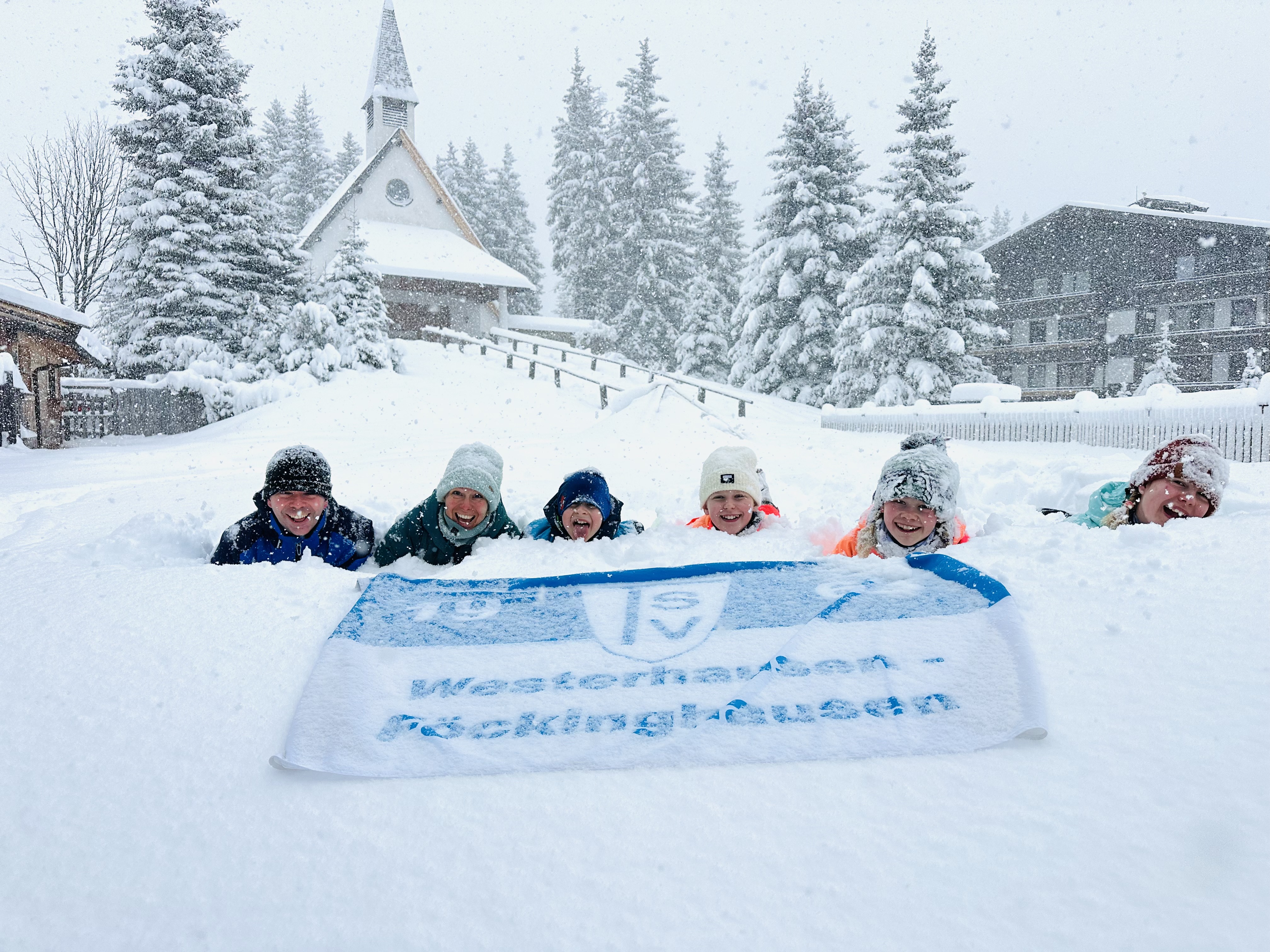 TSV on Tour Familie Berkefeld und Meyer zu Westerhausen Auf dem Bild sieht man eine winterliche Szene mit viel Schnee. Im Vordergrund liegen mehrere Kinder (etwa sechs) nebeneinander im tiefen Schnee und schauen lachend in die Kamera. Sie tragen warme Winterkleidung wie Mützen, Jacken und Schals, was darauf hindeutet, dass es sehr kalt ist und es gerade schneit – die Schneeflocken sind deutlich in der Luft zu erkennen. Vor den Kindern liegt ein teilweise mit Schnee bedecktes Banner oder Handtuch, auf dem man noch den Schriftzug „Westdeutscher Rundfunk“ (WDR) erkennen kann. Im Hintergrund steht eine kleine Kirche mit einem spitzen Turm, die ebenfalls von Schnee bedeckt ist. Rund um die Kirche wachsen hohe, verschneite Tannenbäume. Rechts daneben befindet sich ein größeres Gebäude, vermutlich ein Hotel oder Wohnhaus, das ebenfalls tief im Schnee liegt. Die Stimmung des Bildes wirkt fröhlich und verspielt, da die Kinder trotz der Kälte offensichtlich Spaß im Schnee haben.