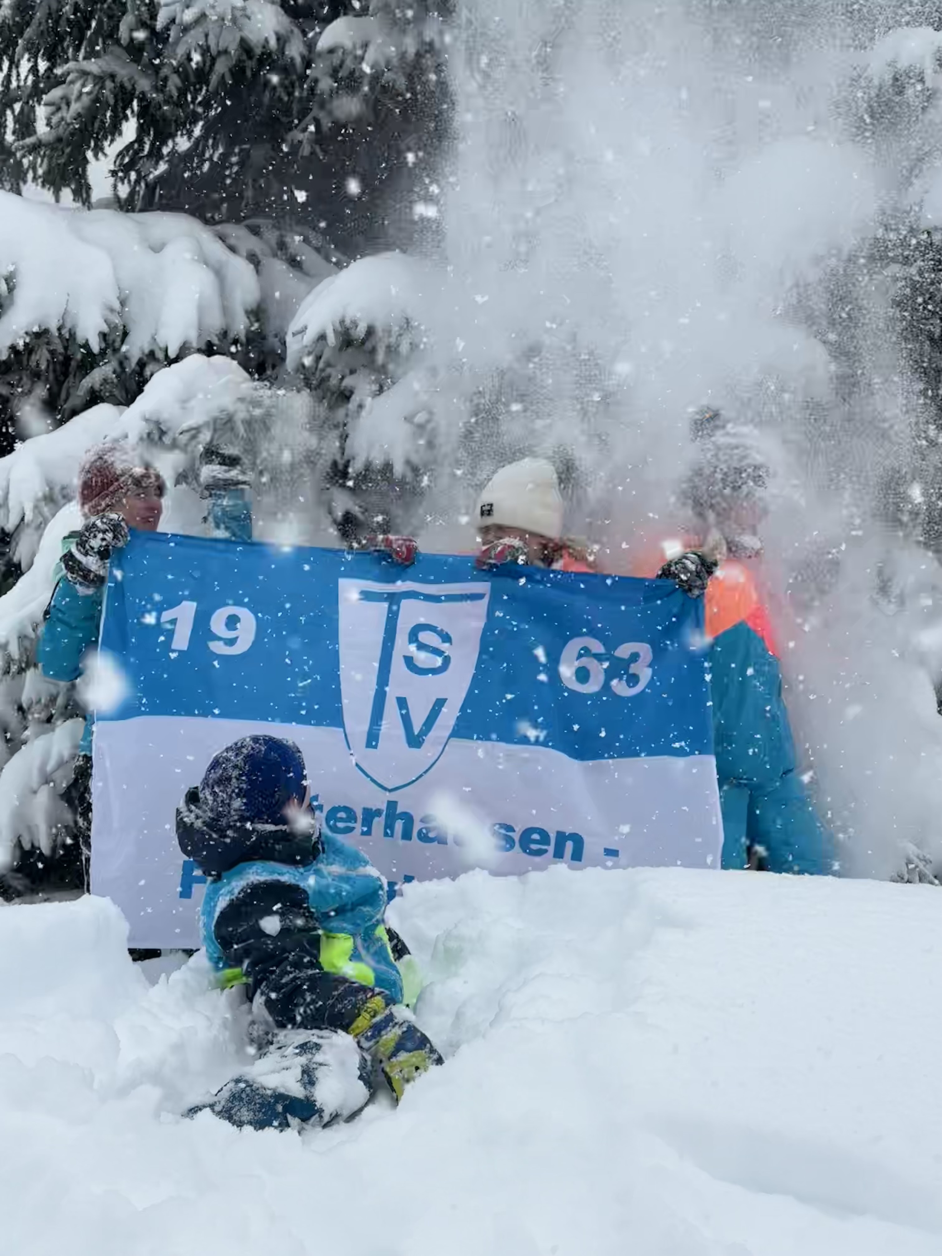 TSV on Tour 2026 Lena, Anna, Piet und Emma Auf dem Bild sieht man eine winterliche Szene mit viel frischem Schnee. Im Vordergrund befindet sich eine Person (vermutlich ein Kind oder Jugendlicher), die bis zu den Knien im tiefen Schnee steht. Die Person trägt warme Winterkleidung, darunter eine Jacke, Handschuhe und eine Mütze. Hinter der Person halten mehrere Menschen ein großes blau-weißes Banner hoch. Auf dem Banner ist deutlich das Logo „TSV“ zu erkennen sowie die Zahlen „1“ und „63“, was vermutlich auf einen Sportverein (z. B. Gründungsjahr 1863) hinweist. Ein besonders dynamischer Moment ist festgehalten: Von oben fällt gerade eine große Menge Schnee herab – möglicherweise wurde er von einem Baum geschüttelt. Der Schnee verteilt sich wie eine Wolke über den Menschen und verdeckt teilweise ihre Gesichter. Im Hintergrund sieht man verschneite Tannenbäume, was die winterliche Atmosphäre verstärkt. Insgesamt wirkt die Szene lebendig und verspielt, als hätten die Beteiligten Spaß daran, im Schnee zu sein und vielleicht gemeinsam eine Aktion oder ein Foto zu machen.