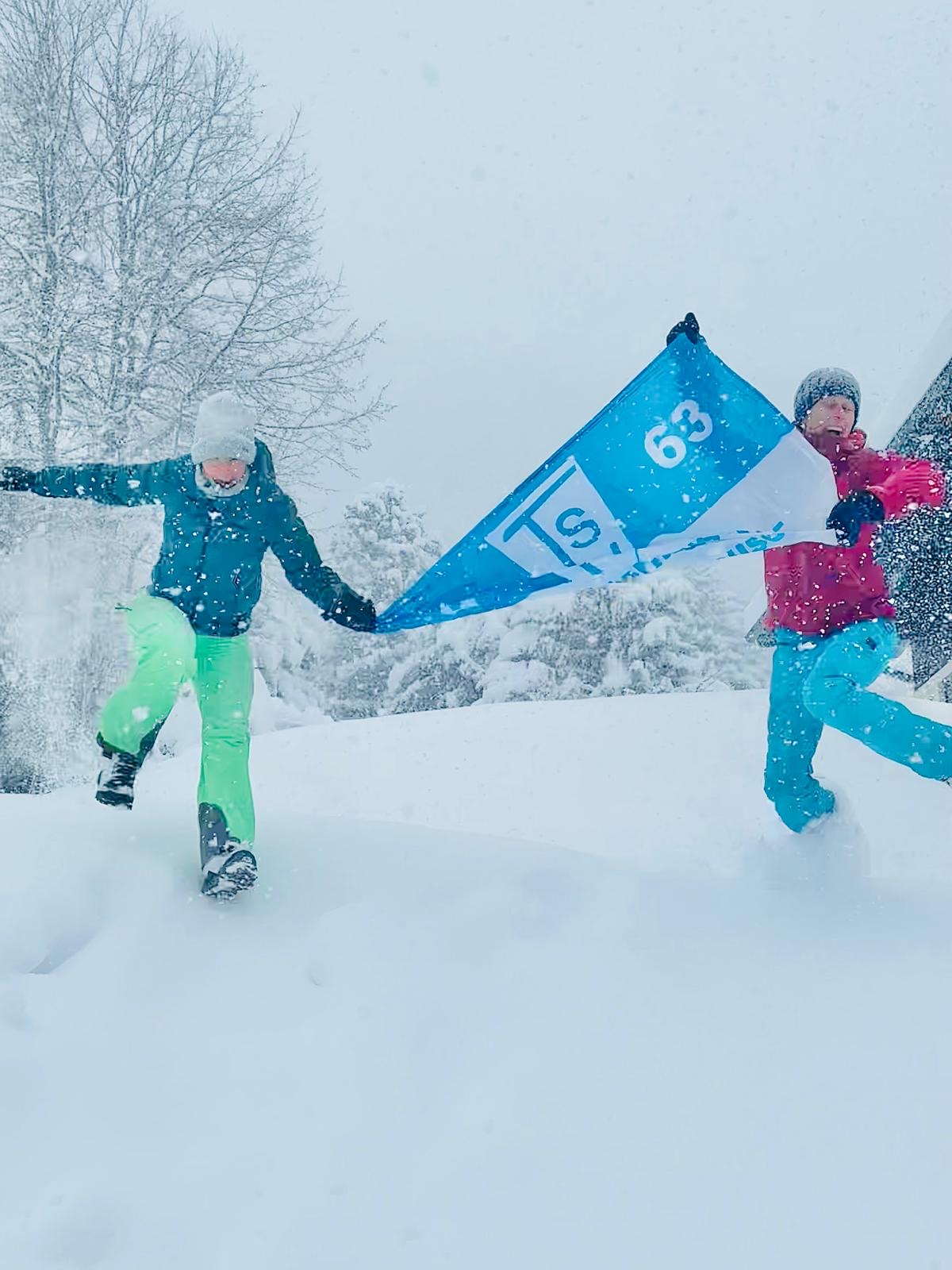 Auf dem Bild sieht man eine verschneite Winterlandschaft mit starkem Schneefall. Der Boden ist von einer dicken Schneeschicht bedeckt, und im Hintergrund stehen kahle, ebenfalls verschneite Bäume.  Im Vordergrund bewegen sich zwei Personen durch den tiefen Schnee. Beide tragen auffällige, warme Winterkleidung:  Die linke Person hat eine dunkle Jacke, eine helle Mütze und eine leuchtend grüne Hose an. Sie hebt ein Bein hoch, als würde sie gerade mühsam durch den tiefen Schnee stapfen oder einen Schritt machen. Die rechte Person trägt eine pinke Jacke, eine blaue Hose und ebenfalls Winteraccessoires wie Handschuhe und Mütze.  Zwischen ihnen halten sie gemeinsam ein blaues Tuch oder Banner, das im Wind oder durch ihre Bewegung leicht gespannt ist.  Die Szene wirkt dynamisch und verspielt – es scheint, als hätten die beiden Spaß daran, sich durch den tiefen Schnee zu bewegen, obwohl die Bedingungen anstrengend sind.