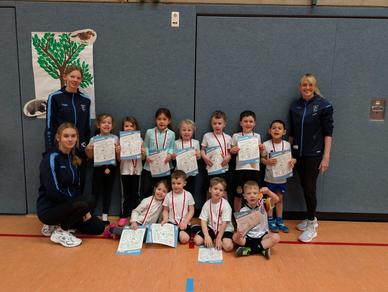 Kinderturnfest am 21.02.2026 Gruppenfoto in einer Sporthalle: Mehrere Kinder stehen und sitzen gemeinsam mit zwei Trainerinnen. Die Kinder tragen Sportkleidung, Medaillen um den Hals und halten Urkunden in den Händen. Alle blicken in die Kamera und wirken stolz und fröhlich.