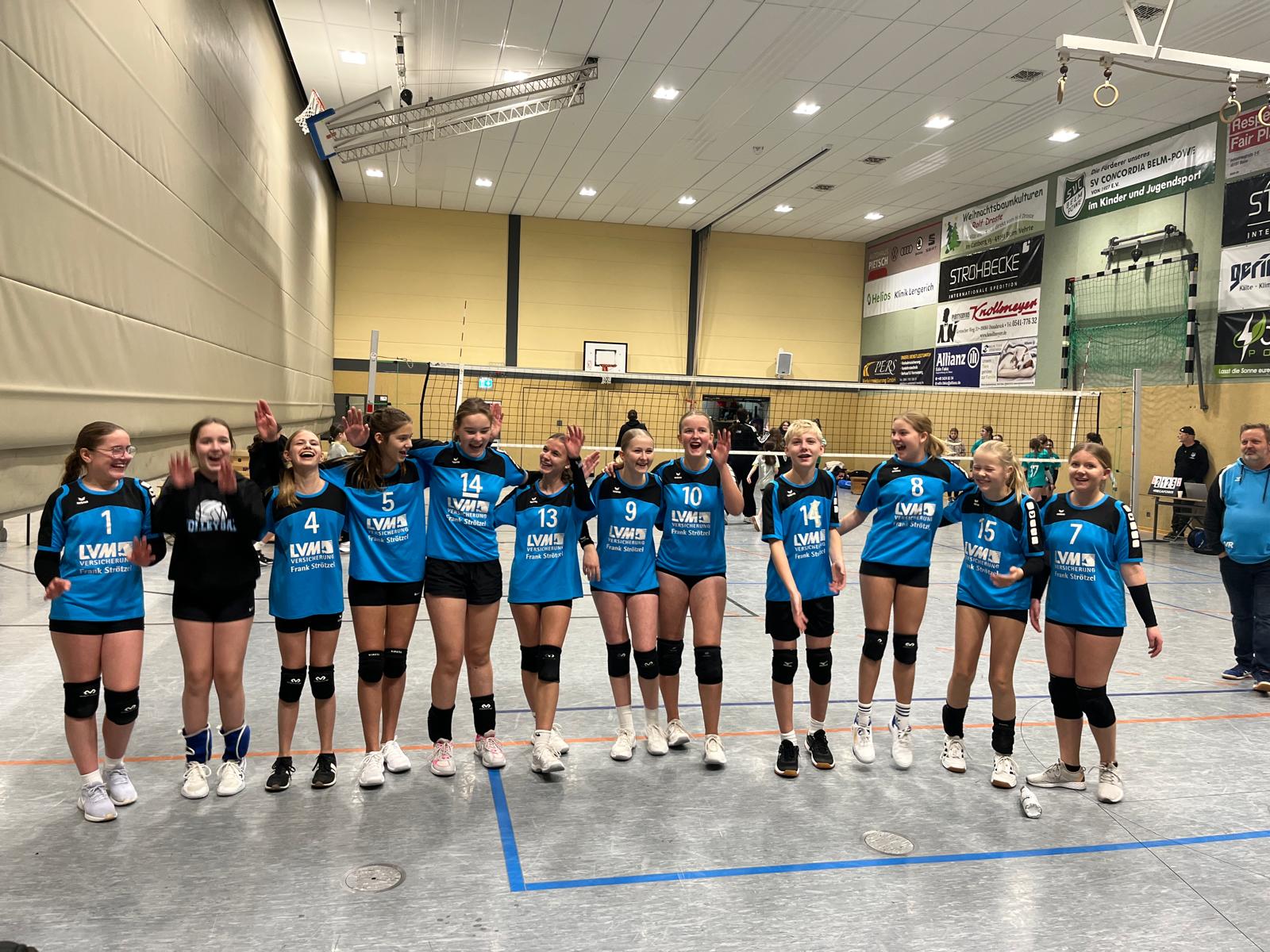 Erster Spieltag Volleyball U16 Auf dem Foto steht eine Volleyball-Mädchenmannschaft in einer Sporthalle. Die Gruppe besteht aus elf Spielerinnen, die nebeneinander in einer Reihe stehen und sich fröhlich präsentieren. Sie tragen überwiegend blaue Trikots mit schwarzen Shorts und Knieschonern. Jede hat eine individuelle Rückennummer auf dem Trikot. Einige heben die Hände, lachen oder schauen sich gegenseitig an – die Stimmung wirkt gelöst, glücklich und voller Teamgeist. Im Hintergrund ist ein Volleyballfeld mit Netz zu sehen, außerdem mehrere Werbebanner an der Hallenwand. Am rechten Bildrand stehen einige Erwachsene, vermutlich Trainer oder Betreuer, die ebenfalls das Geschehen beobachten. Das Gesamtbild vermittelt einen sportlichen, gemeinschaftlichen und motivierten Moment – wahrscheinlich nach einem Spiel oder einem erfolgreichen Wettkampf.