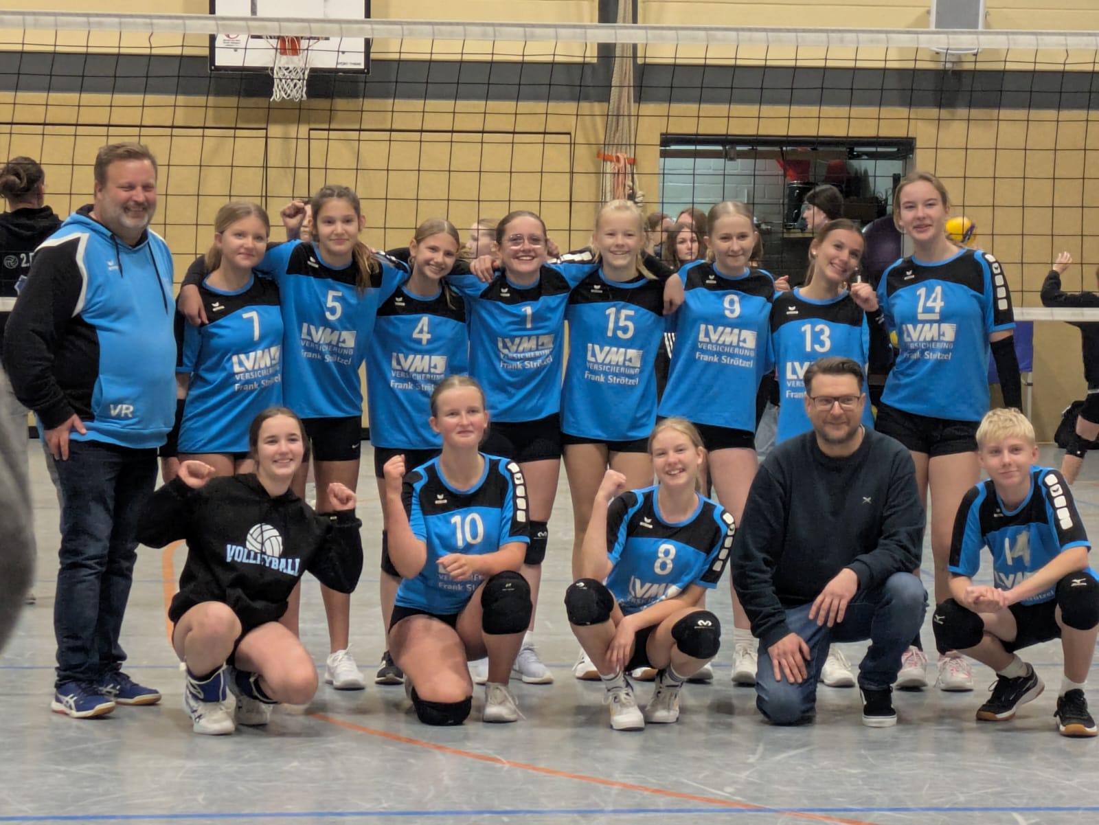 U16 - Volleyball Gruppe von Jugendlichen in blauen Volleyballtrikots mit weißen Nummern und schwarzen Shorts in einer Sporthalle vor einem Volleyballnetz.