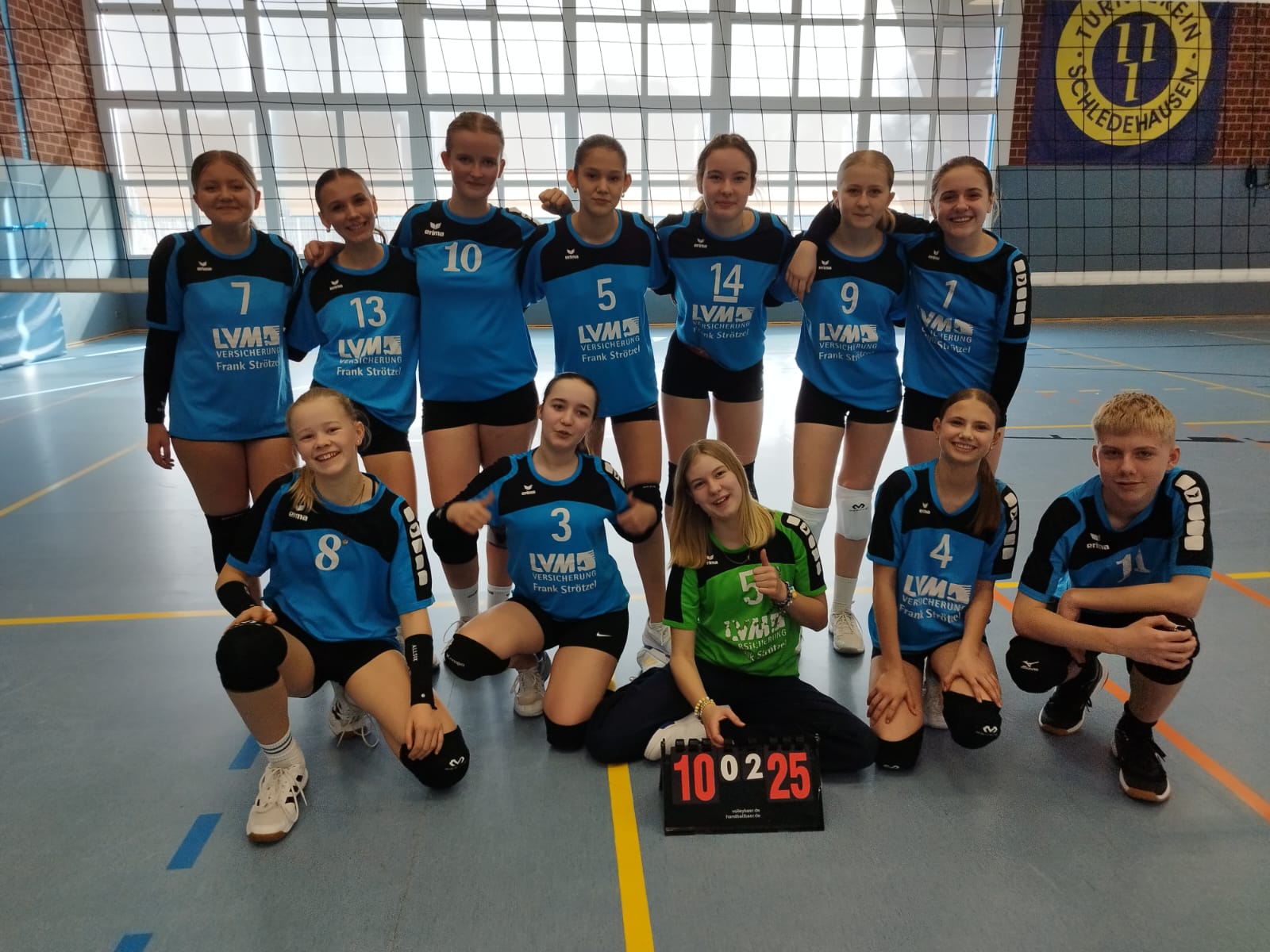 Volleyball U16 - TSV Westerhausen - letzter Spieltag 08.03.2026 Das Bild zeigt eine Jugend-Volleyballmannschaft in einer Sporthalle. 🏐 Vordergrund: Vor dem Team steht eine kleine Anzeigetafel mit dem Ergebnis „10 : 25“. Davor knien mehrere Spielerinnen und ein Spieler. In der Mitte sitzt eine Spielerin im grünen Trikot, vermutlich die Libera, und zeigt mit dem Daumen nach oben. Mitte: Hinter ihnen steht eine Reihe weiterer Spielerinnen. Sie tragen blaue Trikots mit schwarzen Ärmeln und Rückennummern. Auf den Trikots ist ein Sponsor („LVM Versicherung“) zu sehen. Die Spielerinnen haben ihre Arme teilweise umeinander gelegt und lächeln in die Kamera. Hintergrund: Im Hintergrund befindet sich ein Volleyballnetz und eine große Fensterfront der Sporthalle. An der Wand hängt außerdem eine Vereinsfahne des Turnvereins Schledehausen. Gesamteindruck: Das Foto ist ein Mannschaftsbild einer U16-Volleyballmannschaft nach einem Spieltag. Die Gruppe wirkt gut gelaunt und präsentiert gemeinsam das Spielergebnis.