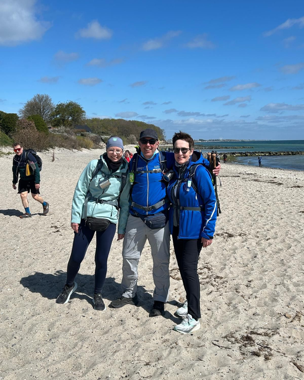 Mega Marsch Ostsee 3d Das Bild zeigt drei Personen, die gemeinsam an einem Strand stehen und in die Kamera lächeln. Sie tragen Outdoor- bzw. Wanderkleidung mit Rucksäcken und wirken, als wären sie gerade unterwegs oder auf einer Tour. In der Mitte steht eine Person mit Sonnenbrille und blauer Jacke, die die beiden anderen freundschaftlich umarmt. Links daneben steht eine Person mit heller Jacke und Mütze, rechts eine weitere Person ebenfalls in blauer Jacke und mit Brille. Der Strand ist hell und sandig, im Hintergrund erstreckt sich das Meer mit ruhigem Wasser und einigen Wellenbrechern aus Steinen. Weiter hinten sind noch andere Spaziergänger zu sehen. Links im Hintergrund wachsen Bäume und Büsche, die auf eine Küstenlandschaft hindeuten. Der Himmel ist strahlend blau mit vereinzelten kleinen Wolken, was auf einen sonnigen, aber möglicherweise windigen Tag schließen lässt. Insgesamt vermittelt das Bild eine fröhliche, aktive Stimmung und erinnert an einen gemeinsamen Ausflug oder eine Wanderung am Meer.