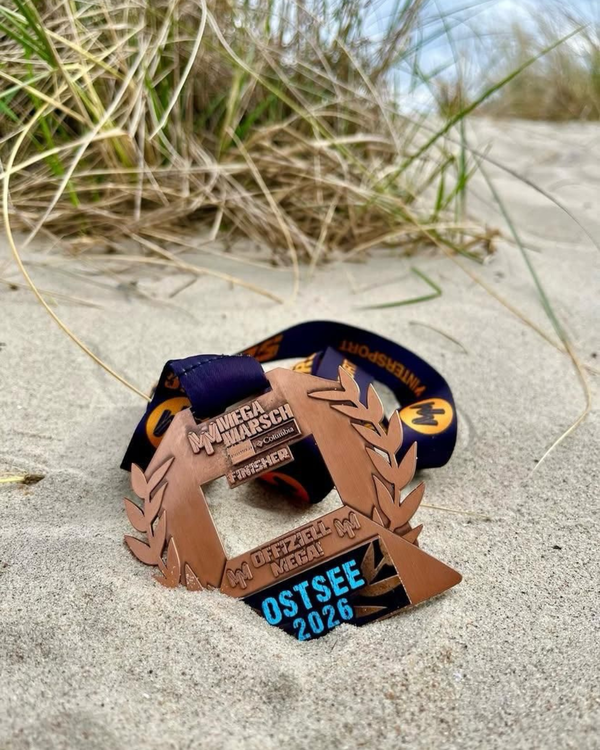 Mega Marsch Ostsee 2 Auf dem Bild ist eine Medaille zu sehen, die auf hellem Sand liegt, vermutlich an einem Strand. Im Hintergrund erkennt man unscharf dünne, trockene Gräser, wie sie oft in Dünen vorkommen. Die Medaille ist bronzefarben und hat ein dekoratives Design mit Lorbeerzweigen an den Seiten. Oben steht „MV Mega Marsch“, darunter kleinere Schrift mit Ortsnamen (u. a. „Columbia“ und „Fischer“ sind angedeutet). In der Mitte ist eine ausgesparte Form, und unten ist ein schwarzes Element mit türkisfarbener Schrift, auf dem „OSTSEE 2026“ steht. Außerdem ist „M. Oeffzell MV Mega“ (teilweise schwer lesbar) eingraviert. An der Medaille ist ein dunkles Band befestigt, das im Sand liegt. Insgesamt wirkt das Bild wie eine Nahaufnahme einer Finisher- oder Eventmedaille, die an einem Ostseestrand fotografiert wurde.