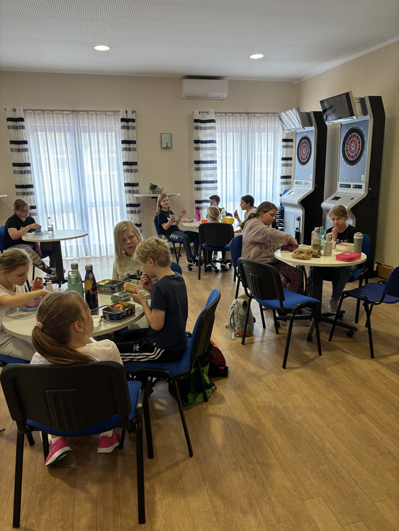 Frühstück Feriencamp Das Bild zeigt einen hellen Aufenthaltsraum, in dem mehrere Kinder an kleinen runden Tischen sitzen und gemeinsam essen. Die Kinder haben Brotdosen, Trinkflaschen und verschiedene Snacks vor sich stehen. Einige unterhalten sich miteinander, während andere sich auf ihr Essen konzentrieren. Der Raum wirkt freundlich und ordentlich. Große Fenster mit hellen Vorhängen lassen viel Tageslicht herein. Die Möbel bestehen aus einfachen Tischen und Stühlen, und der Boden ist aus Holz. An der Wand im Hintergrund stehen zwei elektronische Dartautomaten, was darauf hindeutet, dass der Raum auch zur Freizeitgestaltung genutzt wird. Die Kinder tragen bequeme Alltagskleidung, und insgesamt entsteht der Eindruck einer Pause, zum Beispiel während eines Ausflugs oder eines Schultages. Die Atmosphäre ist entspannt und ruhig.