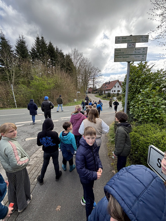Schnitzeljagd Feriencamp Das Bild zeigt eine Gruppe von Kindern und einigen Erwachsenen, die gemeinsam an einer Straße entlanggehen. Es wirkt wie ein Ausflug oder ein Spaziergang, möglicherweise von einer Schulklasse oder einer Gruppe. Die Kinder tragen überwiegend Jacken und warme Kleidung, was darauf hindeutet, dass es eher kühl ist. Einige schauen nach vorne, während andere sich umdrehen oder miteinander sprechen. Die Gruppe befindet sich an einer Straßenkreuzung in einer ländlichen oder kleinstädtischen Umgebung. Rechts im Bild steht ein Wegweiser mit Entfernungsangaben zu nahegelegenen Orten. Im Hintergrund sind Wohnhäuser mit roten Dächern sowie Bäume und Büsche zu sehen. Links säumen hohe, dichte Bäume den Straßenrand. Der Himmel ist stark bewölkt mit dunklen Wolken, was eine leicht trübe Stimmung erzeugt. Insgesamt vermittelt das Bild den Eindruck eines organisierten Spaziergangs oder einer Wanderung in einer ruhigen Gegend.