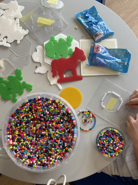 Feriencamp Bügelperlen Auf dem Bild sieht man einen runden, hellen Tisch, auf dem Bastelmaterialien ausgebreitet sind. Im Mittelpunkt steht eine große Schüssel voller bunter, kleiner Steckperlen in vielen verschiedenen Farben. Daneben befindet sich ein kleineres Gefäß, ebenfalls mit solchen Perlen gefüllt. Auf dem Tisch liegen mehrere transparente Steckplatten mit unterschiedlichen Formen, darunter Tiere wie Pferde oder andere Figuren. Einige dieser Formen sind bereits mit Perlen in Farben wie Grün, Rot oder Weiß teilweise ausgefüllt. Besonders auffällig ist eine rote Pferdefigur sowie eine grüne Form daneben. Außerdem sind zwei blaue Verpackungen zu sehen, vermutlich von den Perlen oder Zubehör. In der Nähe liegt ein rundes, gelbes Steckbrett. Rechts im Bild erkennt man die Hände einer Person, die gerade dabei ist, Perlen auf eine transparente Steckplatte zu stecken und ein Muster zu legen. Insgesamt vermittelt das Bild eine kreative Bastelsituation, vermutlich von einem Kind oder einer Person, die gerade mit Bügelperlen arbeitet.