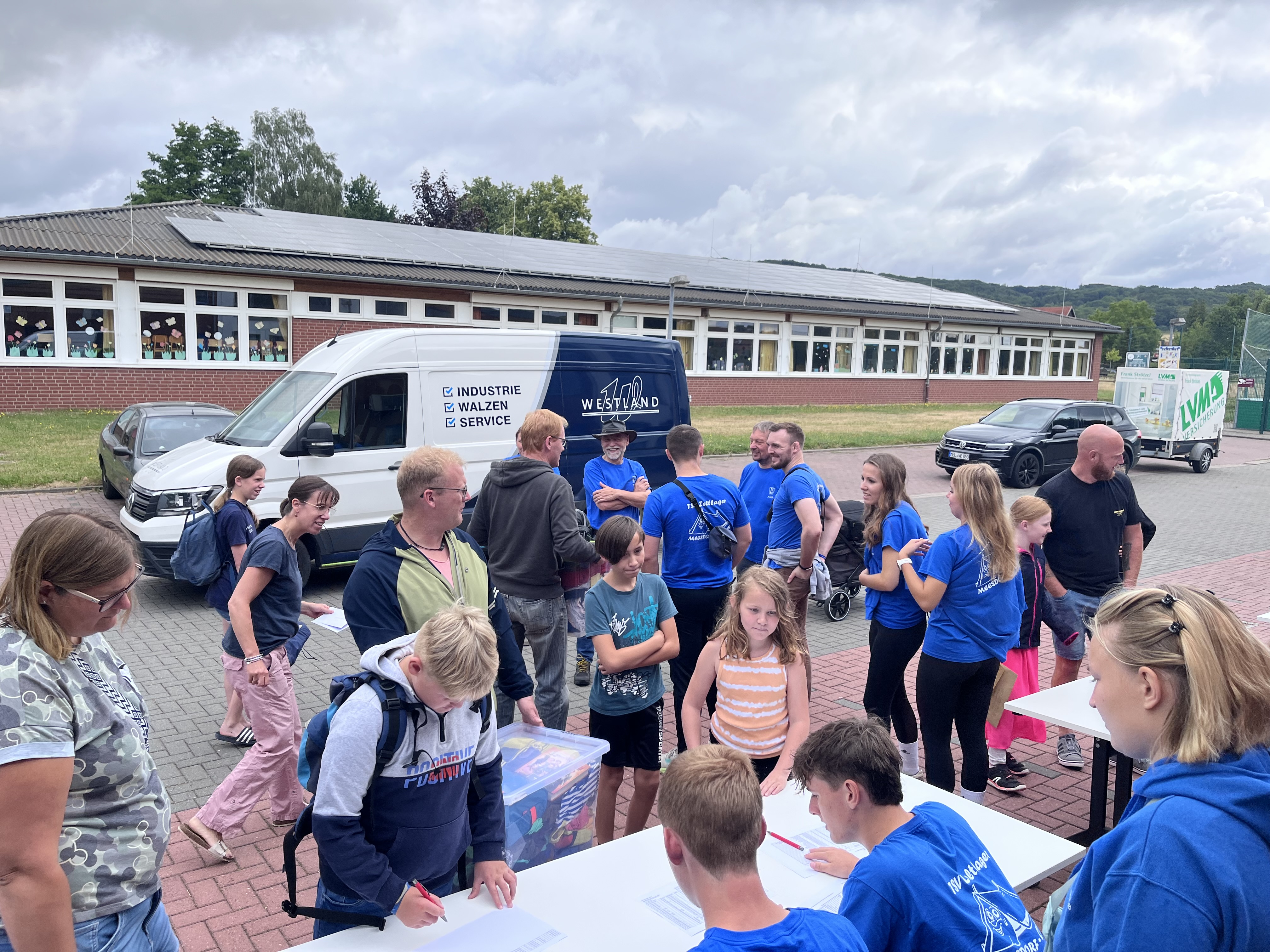 Zeltlager Meesdorf 2026 - Tag 1 Check -In Gruppe von Kindern und Jugendlichen steht auf einem Schulhof, einige schreiben an einem weißen Tisch, im Hintergrund ein weißer Transporter mit Aufschrift und ein Schulgebäude