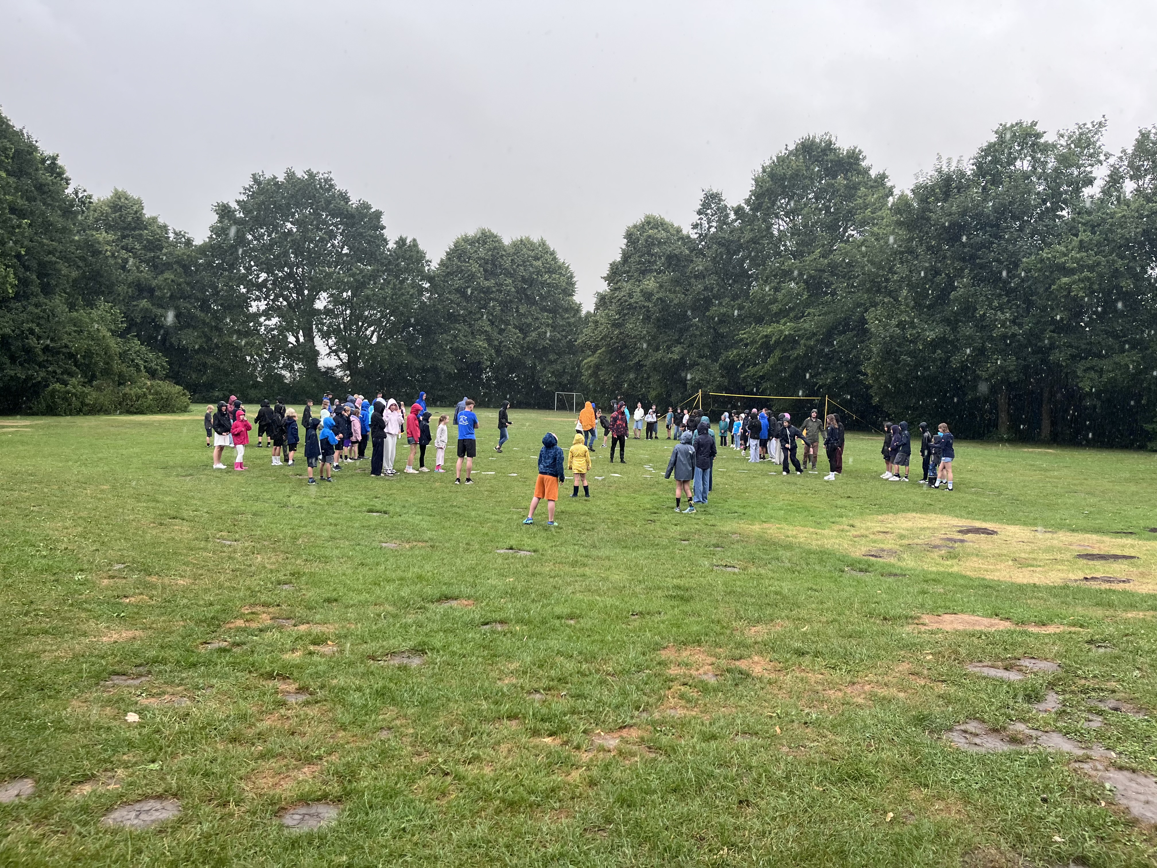 Zeltlager Meesdorf 2026 - Tag 1 - Ankommen auf dem Zeltplatz Gruppe von Kindern und Jugendlichen steht verteilt auf einer großen Wiese vor Baumreihe bei bewölktem Himmel