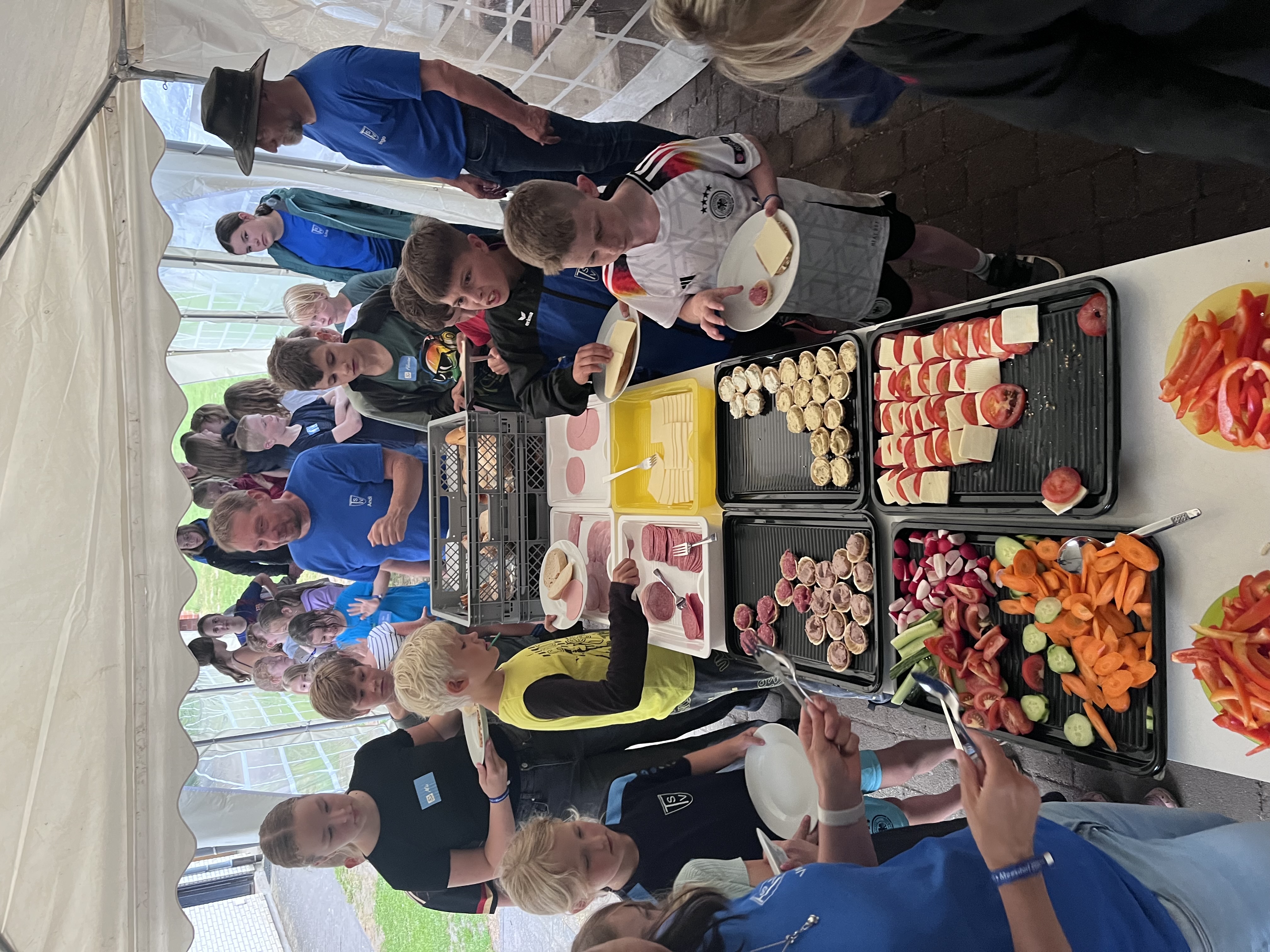 Zeltlager Meesdorf 2026 - Tag 1 - Abendessen Kinder und Erwachsene stehen unter einem Zelt an einem langen Tisch mit Tellern und Tabletts voller Wurst, Käse, Gemüse und Brötchen